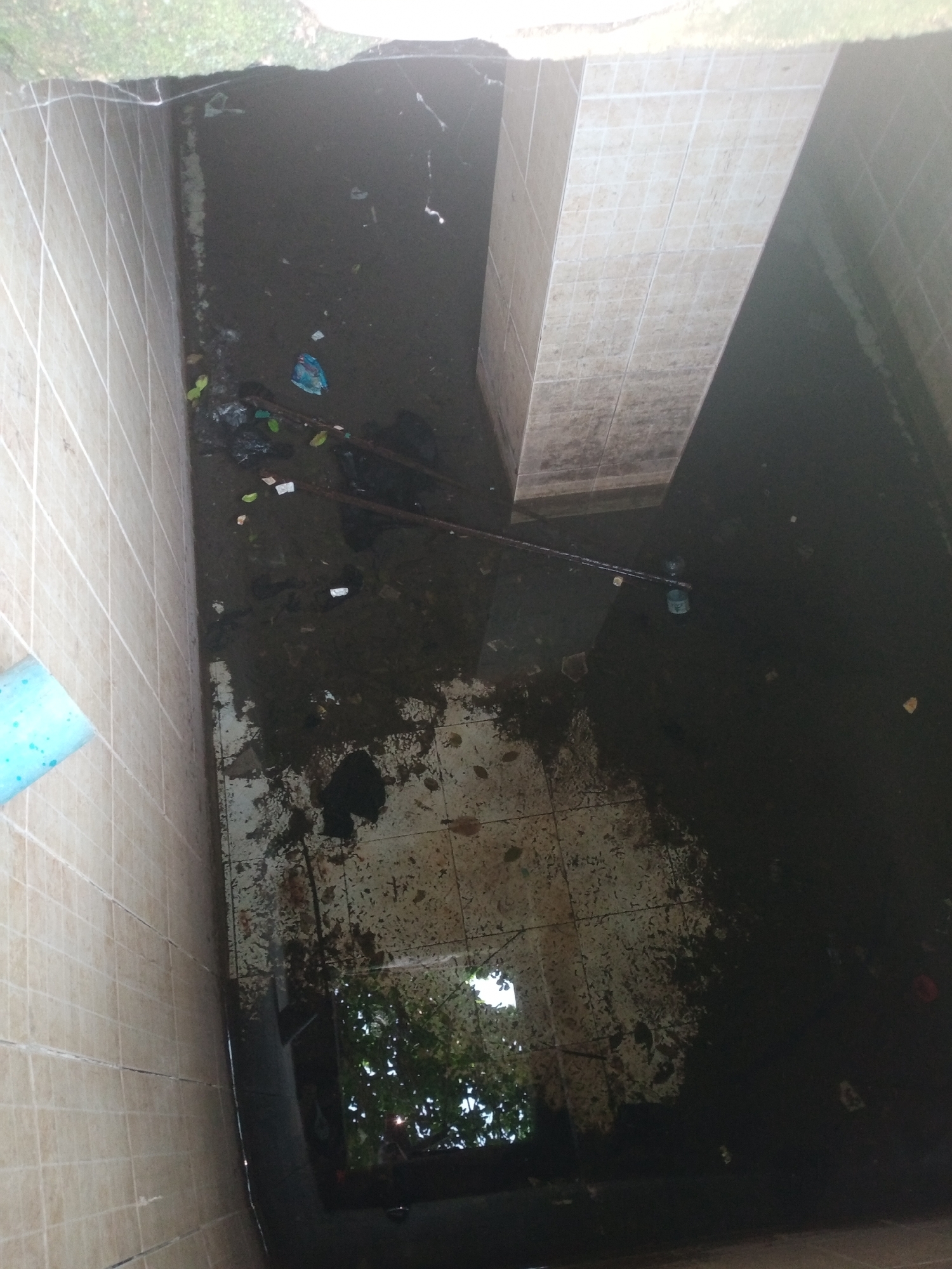 The image shows a neglected, flooded area with debris. The floor is tiled and appears to be submerged in murky water, with trash, leaves, and other detritus scattered throughout. A tiled structure juts into the frame from the right. The walls on the left are also tiled. The scene gives the impression of an outdoor space that has fallen into disrepair and is experiencing water accumulation, possibly due to poor drainage. The setting could be a courtyard, a drainage channel, or a similar urban outdoor space in Jalingo, Nigeria.