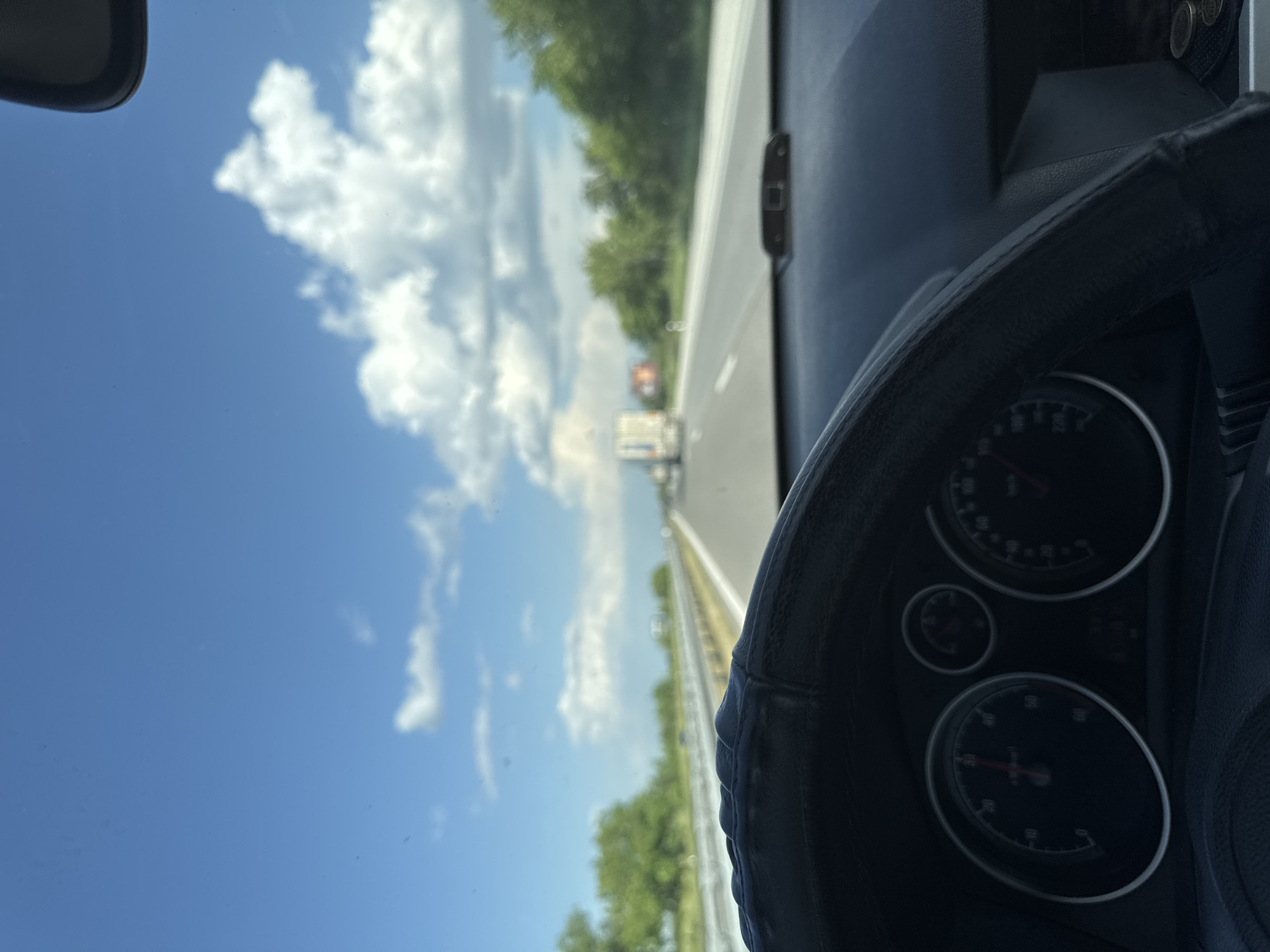 The photograph shows an interior view from a vehicle, likely a car, looking out onto a highway with a bright blue sky and scattered white clouds. A steering wheel is visible in the foreground, obscuring part of the dashboard. The highway is bordered by greenery on either side, suggesting a rural or suburban setting. In the distance, vehicles can be seen on the road. There is no information to determine if the location is in the city of Shtarkovo, Bulgaria.