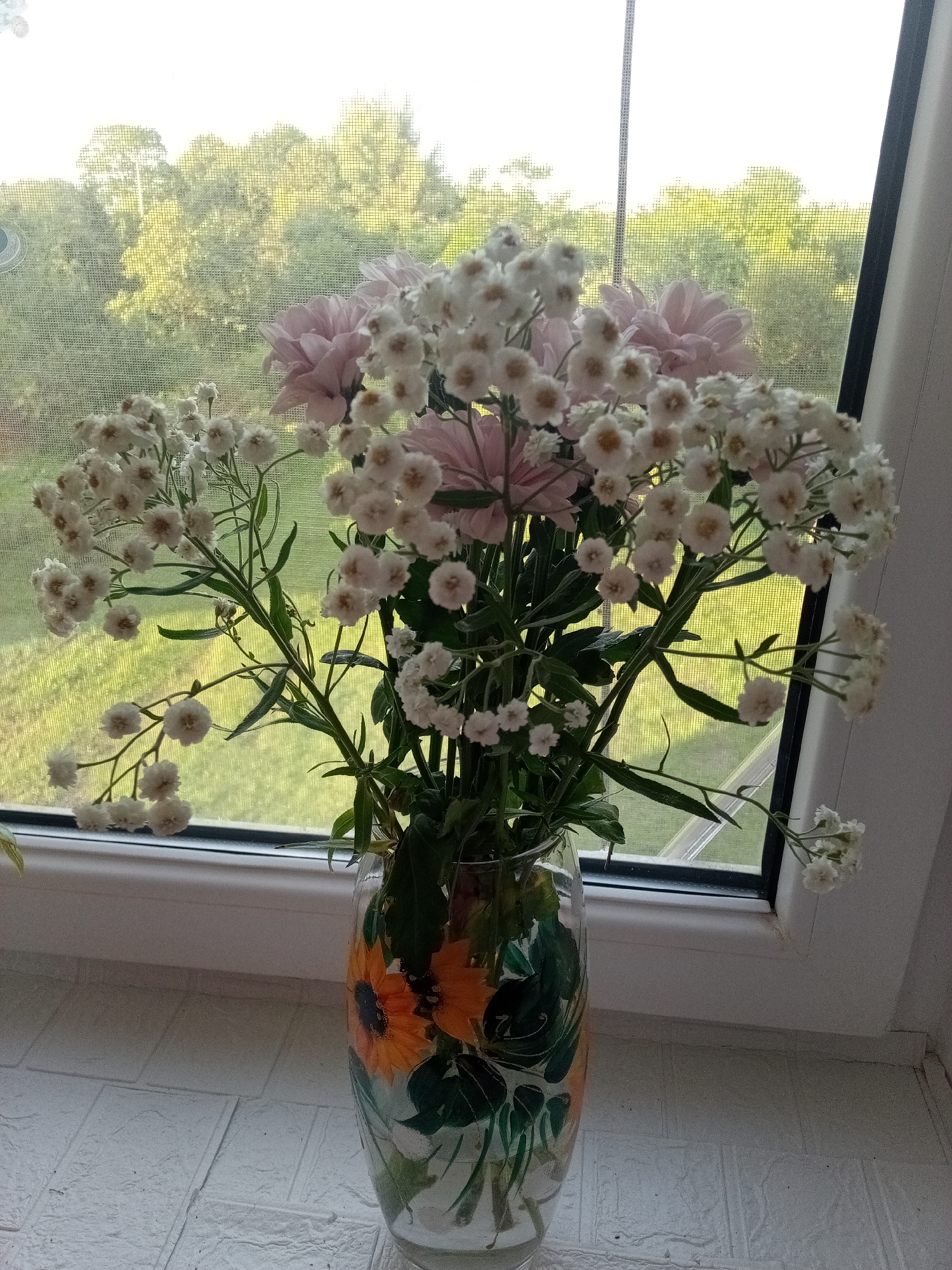 A vase of flowers sits on a windowsill, with a view of trees and greenery through the window. The vase is clear glass and decorated with painted sunflowers and leaves. The bouquet consists of pinkish-purple flowers and small white flowers, possibly baby's breath, with green foliage. The flowers appear fresh and are arranged in a loosely held bunch. The windowsill is white and textured, resembling a brick pattern. Through the window, a sunny day is implied by the bright, slightly washed-out light and the green of the foliage. A window screen is visible, obscuring some of the detail of the outdoor scene. The overall impression is one of domestic tranquility and a connection to nature. The location context of Chernyshevskoye, Russia, suggests a rural or suburban setting. No people are visible in the image. There is no visible text. The time of day is likely daytime, given the brightness of the scene.