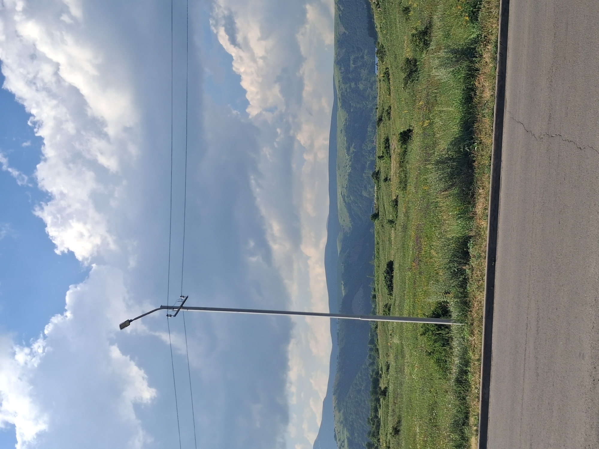 The image shows a wide, paved road in the foreground with a distinct crack running across it. Beyond the road is a grassy field dotted with scattered bushes. A tall, slender streetlight stands prominently in the field, its pole angled slightly. In the background, rolling green hills form a natural landscape under a sky filled with dramatic, fluffy white clouds and patches of blue. The scene appears to be an outdoor setting in Jermuk, Armenia, possibly along a roadside with views of the surrounding countryside. There are no people or animals visible in the image. The overall impression is one of a serene, natural landscape with man-made elements like the road and streetlight.