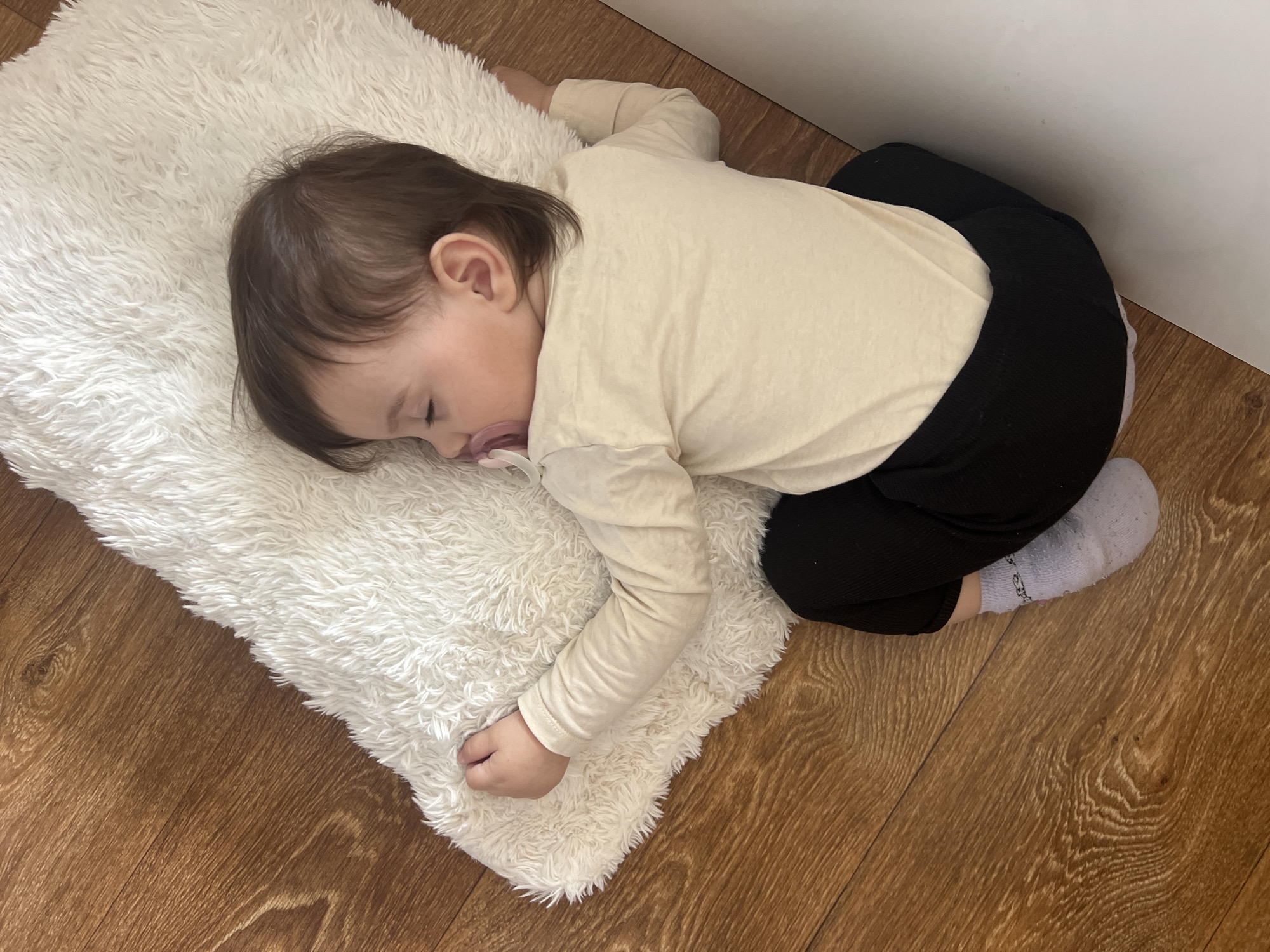 A young child sleeps on a white fluffy rug on a brown laminate floor. The child is wearing a light beige long-sleeved shirt and black pants with a single grey sock on one foot. The child has dark hair and skin, and a pacifier is visible in the child's mouth. The rug is positioned near a white wall in the corner of the room. The location of Elena, Bulgaria is not discernable from the image.
