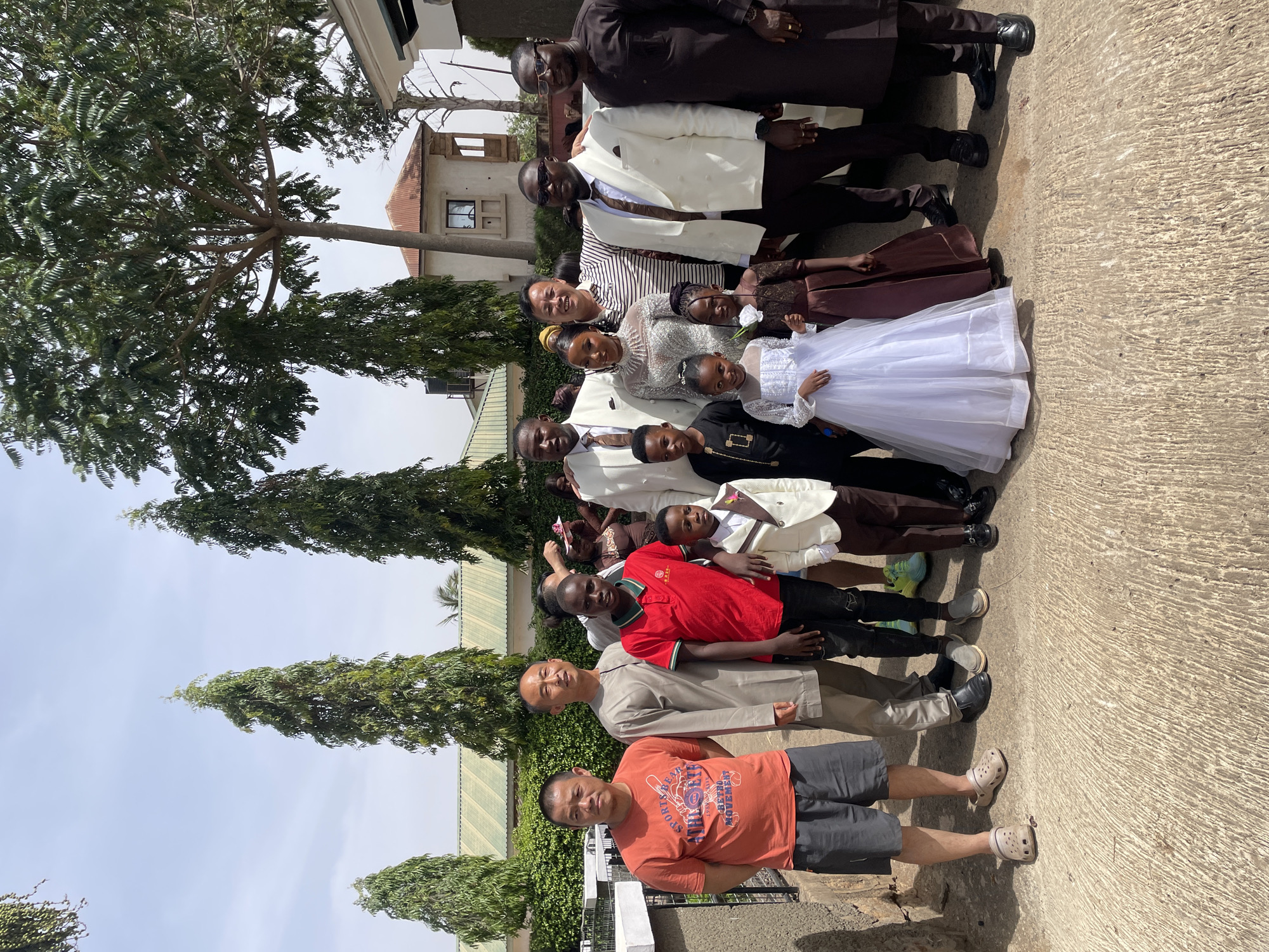 A group of approximately 13 people of varying ages are standing together outside. The group consists of adults and children, some of whom are dressed in formal attire, including white dresses and suits. Several people are wearing casual clothing. The group appears to be posing for a picture.
The scene is set outdoors on a paved surface, likely a street or walkway. Buildings and trees are visible in the background. Based on the appearance of the buildings and vegetation, and the user input, the location is likely in the city of Jos, Nigeria.