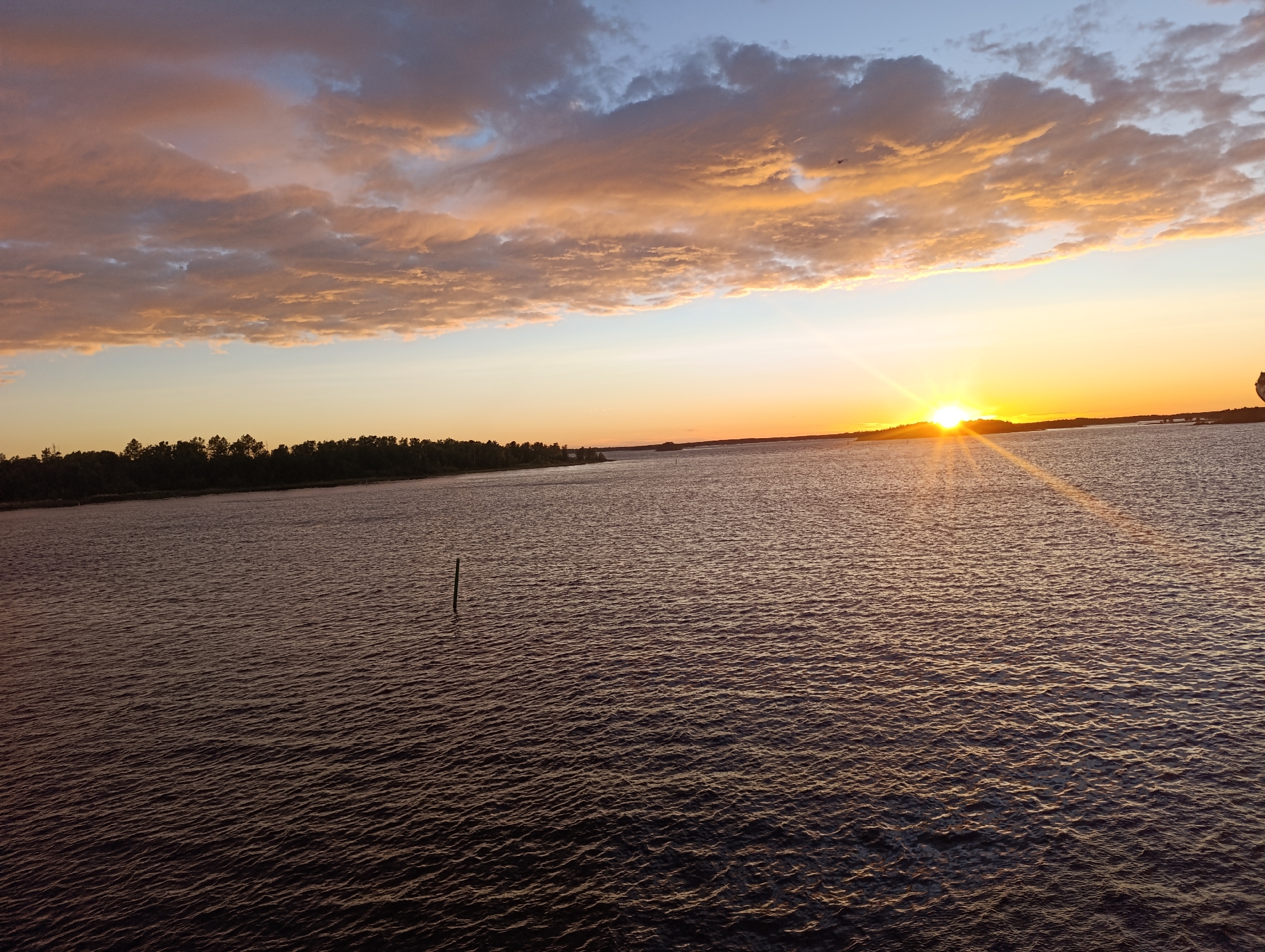 A wide-angle photograph captures a sunset over a body of water, likely the sea or a large lake, in Jakobstad, Finland. The sky is filled with dramatic clouds, tinged with hues of orange, pink, and blue. The sun, a bright disc of light, is setting near the horizon, casting long rays of golden light across the water. The water itself is a deep, dark blue, with ripples reflecting the sky's colors. A dark line of trees is visible along the distant shoreline, and a single, thin wooden post stands in the water in the foreground, breaking the surface. The overall impression is one of natural beauty and tranquility.