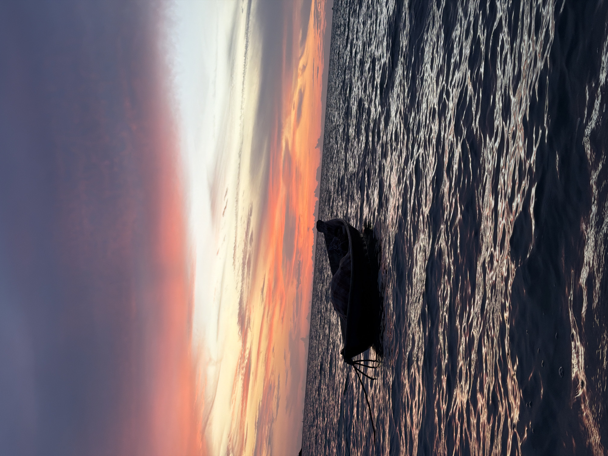 A small, dark boat is anchored in the calm, dark water of the ocean. The boat is silhouetted against a dramatic sunset sky. The sky is filled with vibrant colors, ranging from deep purple and pink at the top to bright orange and yellow closer to the horizon. Wispy clouds stretch across the sky, catching the light of the setting sun. The water reflects the colors of the sky, creating a shimmering effect. The scene appears to be captured during twilight on the coast of Ko Samui District, Thailand. There are no people visible in the image.