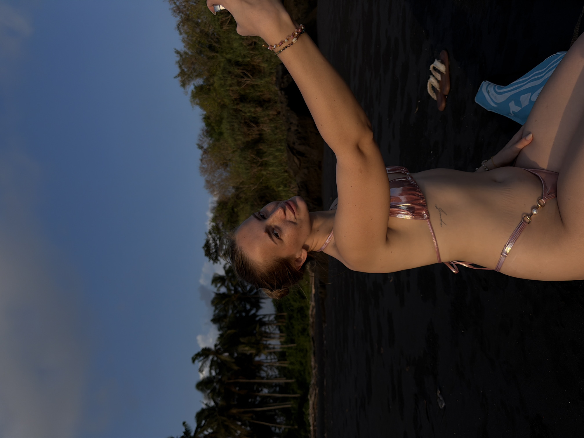 A woman in a pink bikini takes a selfie at the beach. She is lying down on black sand, with palm trees and lush greenery in the background. The sky is a clear blue. A pair of flip-flops and a blue towel are visible on the sand next to her. The lighting suggests it is either early morning or late afternoon. The woman appears relaxed and happy. There is no visible text in the image.