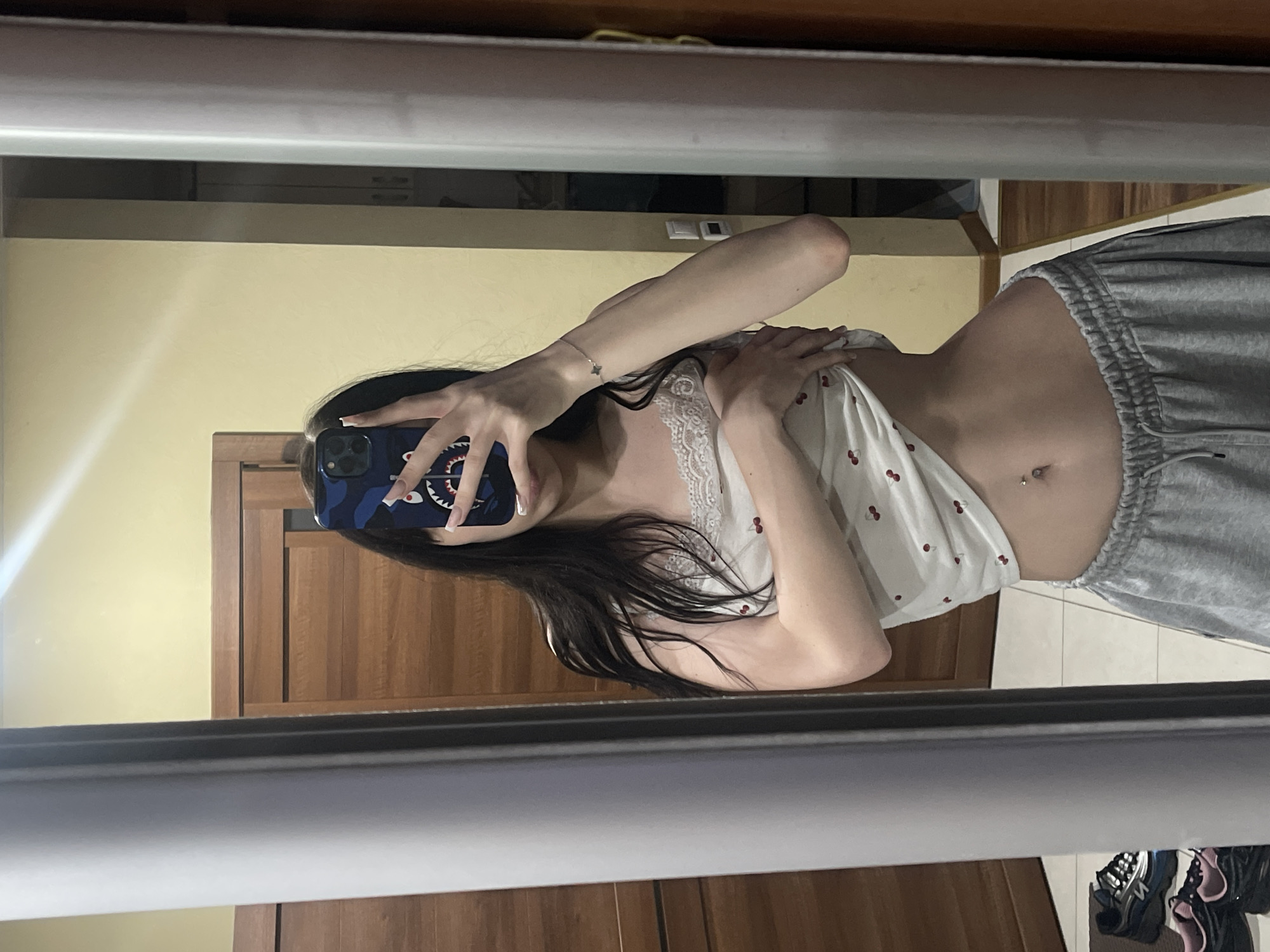 A young woman with long, dark hair is standing in front of a mirror in what appears to be a bedroom. She is wearing a white camisole with a cherry print and grey sweatpants. The woman is holding her phone up to take a selfie, and is lifting the hem of her camisole to expose her stomach, which has a belly button piercing. The room has wooden doors and pale yellow walls. Shoes are visible on the floor in the reflection of the mirror. The location is the city of Khimki, Russia.