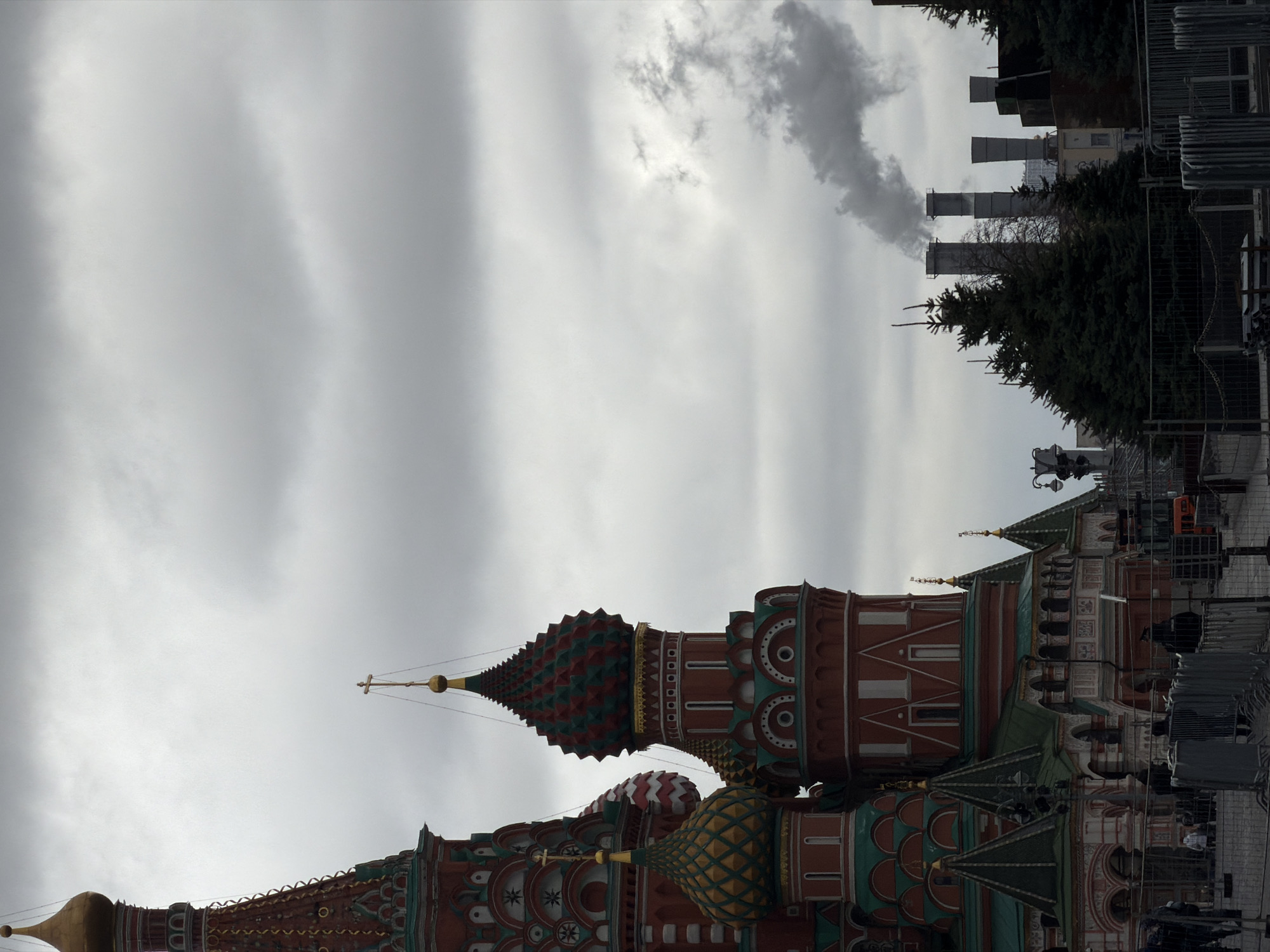 The image depicts a low-angle view of St. Basil's Cathedral in Moscow, Russia, set against a cloudy sky. The Cathedral's distinctive onion domes, colored in shades of red, green, gold, and brown, are prominently featured. In the distance, chimneys emit smoke into the overcast sky. In the background, there are some fir trees and a building.