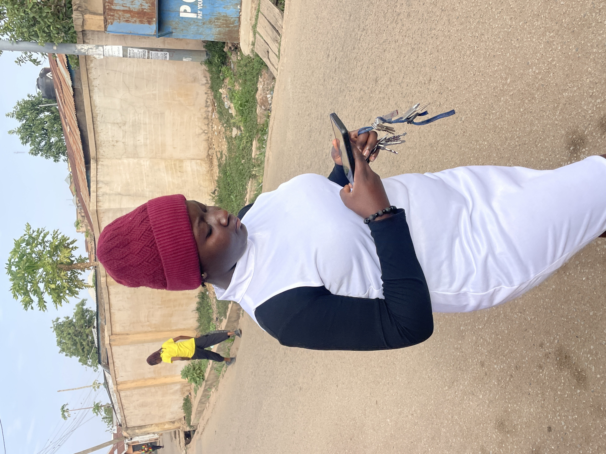 The image, rotated 90 degrees clockwise, captures a street scene likely in Jos, Nigeria, during the daytime. In the foreground, a person is shown in profile, wearing a maroon knitted beanie, a white sleeveless top over a black long-sleeved shirt, and a black beaded bracelet on their right wrist. They are intently looking down at a black smartphone held in both hands, while also holding a large bunch of keys attached to a blue ribbon or lanyard. Their expression appears focused and neutral.
The setting is a paved road with a light brown, speckled surface. Along the side of the road, various structures are visible, including buildings with unpainted concrete walls and rusted corrugated metal roofs. Green trees and scattered vegetation grow along the roadside and behind the buildings. In the mid-ground, another person wearing a yellow t-shirt and dark trousers is seen walking away from the camera.
In the upper right portion of the background, a blue metal utility box or signpost is visible, featuring the partially legible text "PAY YOU." Other posters with unreadable text are affixed to a pole near the blue structure. The lighting is bright and diffused, suggesting either an overcast sky or soft daylight without harsh shadows. The scene depicts an ordinary moment in an urban or semi-urban environment.