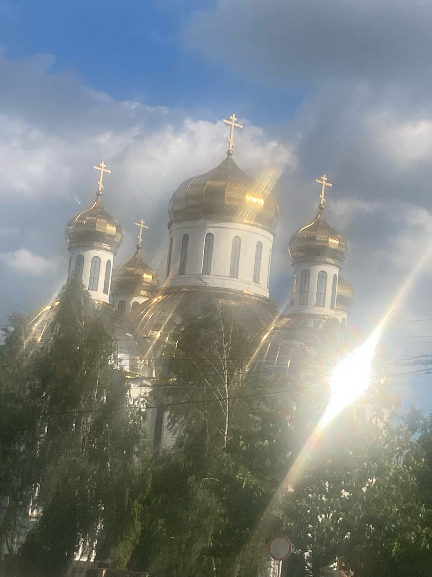 The image shows the golden domes of a church, possibly the Church of St. Nicholas, in the city of Brest, Belarus. The church has multiple domes, topped with crosses, and is surrounded by trees. The sun is shining brightly, creating a lens flare across the image. The sky is partly cloudy.