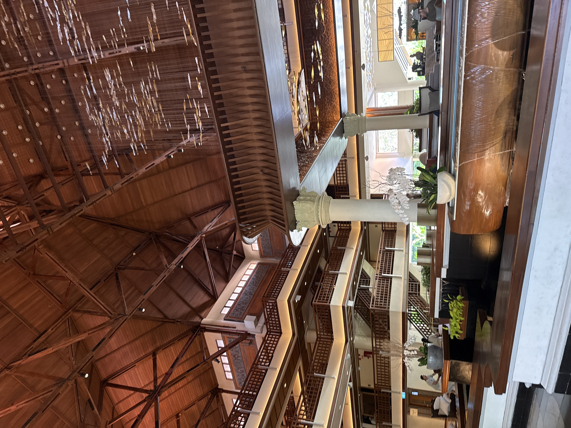 The image captures a wide-angle view of the interior of a grand lobby, likely in a resort or hotel. The architecture features a prominent wooden beam ceiling with intricate structural support, indicative of a tropical design. Multiple levels of balconies with wooden railings are visible, connected by staircases. A modern water feature with cascading water runs across the foreground, bordered by dark wood and white structures. In the background, there are glimpses of people, possibly guests or staff, engaged in activities such as sitting, walking, and interacting. One person is visible near a reception desk. The lighting appears to be a mix of natural light filtering from large windows and artificial ambient lighting, highlighting the warm tones of the wood and creating a serene atmosphere. There's a large floral arrangement with white orchids on a dark pedestal in the mid-ground. Hanging from the ceiling is a decorative mobile or chandelier composed of many small, reflective elements. The overall impression is one of spaciousness, natural materials, and a luxurious, calming environment, consistent with a high-end hospitality establishment in a tropical setting like Kuta Selatan, Indonesia.