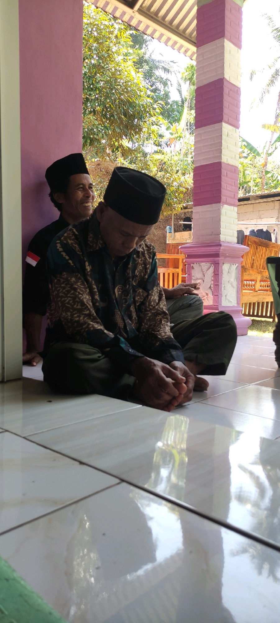 Two men, both wearing black peci hats, are seated on a tiled floor outdoors. The man in the foreground is looking down and appears to be resting his hands on his lap. He is wearing a patterned shirt and green pants. The man in the background is leaning against a pink wall and smiling. He is wearing a black shirt with a white and red Indonesian flag emblem on the sleeve. The scene appears to be in a residential setting in Plunjaran, Indonesia, with a covered porch area with decorative columns in the background, and lush greenery visible beyond. Sunlight reflects off the polished floor.