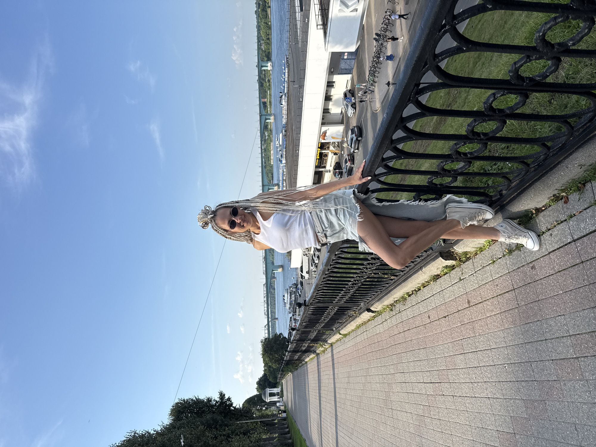 A full-shot, outdoor, eye-level photograph shows a woman standing on a promenade next to a decorative black metal fence. She is positioned in the right half of the frame, leaning on the fence with her right hand. She wears a white tank top, distressed denim shorts, and white high-top sneakers. Her hair is styled in numerous long, platinum blonde braids, and she wears dark oval sunglasses. Behind her, a wide river stretches across the frame, with a green bridge in the distance. Boats are visible on the water, and a white building with a flat roof is situated on the riverbank. Cars are parked in front of the building, and a row of bicycles is chained nearby. To the left of the woman, a paved promenade extends into the distance, lined with trees on its left side. A small white gazebo with a dome is visible further down the promenade. The sky is clear blue with a few wispy clouds. The scene appears to be a sunny day in the city of Rusanovo, Russia.