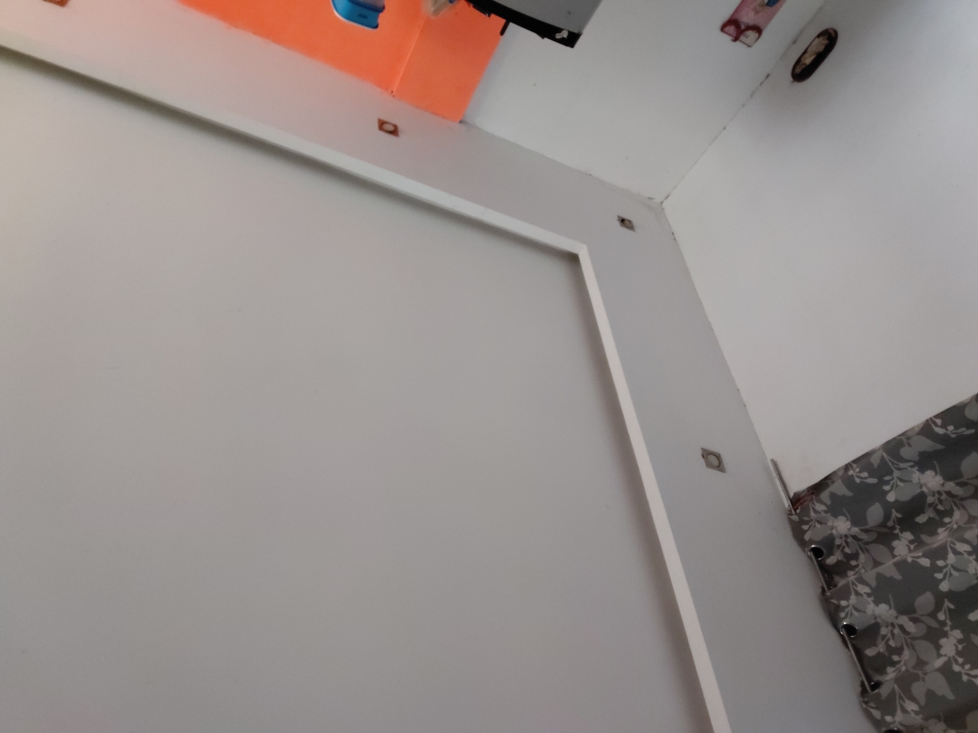 The image captures an upward-angled view of an indoor corner, likely a room. The dominant feature is a light-colored wall (possibly white or light grey) on the left and center, adorned with a rectangular raised molding or frame. This wall meets another white wall at an angle on the right, forming an interior corner of the room.
In the upper left portion, an orange surface or object is visible, partially obscured, with a small blue object next to it. Several small, square-shaped cutouts or openings are present on the white walls near the ceiling, some appearing as dark holes, while one shows a light-colored cover with a dark interior. A patterned curtain, grey with a white floral or leaf design, hangs on the right side of the image, secured with visible grommets.
Towards the upper right, a dark, oval-shaped object (possibly a light fixture or vent) is mounted on the wall, and a smaller, reddish-pink object is also visible nearby. A faint crack runs along the seam where the wall meets the ceiling in the upper right quadrant. No people are visible, and no specific activity is taking place. The time of day and weather are not discernible from the indoor setting, and there is no visible text. The overall impression is a static view of a residential interior in Santa Ana, Venezuela.