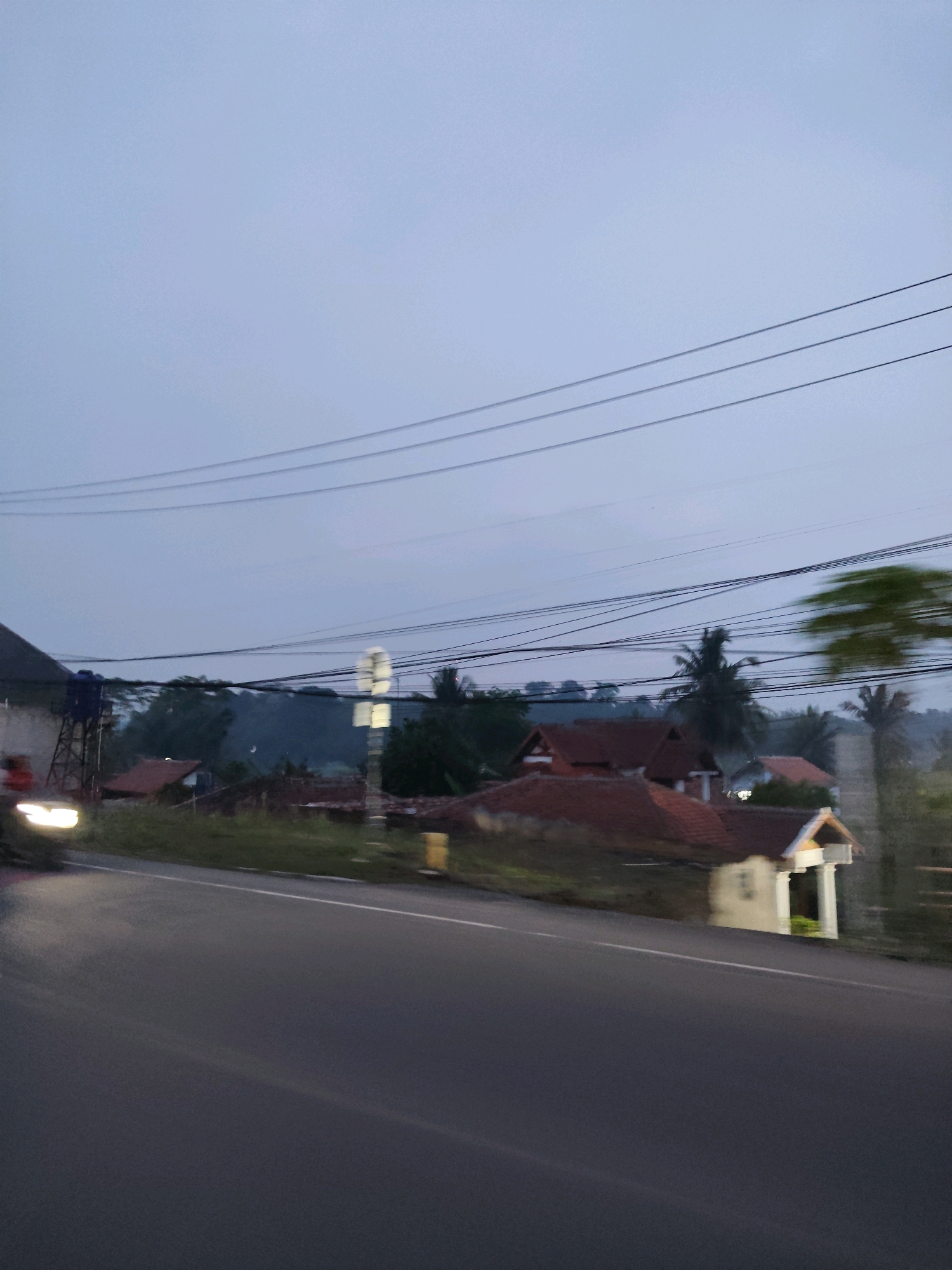 The image depicts a street scene at dusk in Warunggunung, Indonesia. A paved road occupies the foreground, curving to the right. Beyond the road, there are several houses with red-tiled roofs and some vegetation. A structure with a blue water tank on a metal frame is visible on the left side, along with the headlights of a vehicle. Overhead, multiple power lines stretch across the frame. The sky is a pale, overcast blue, suggesting the end of the day. The overall impression is of a quiet, semi-rural residential area.
