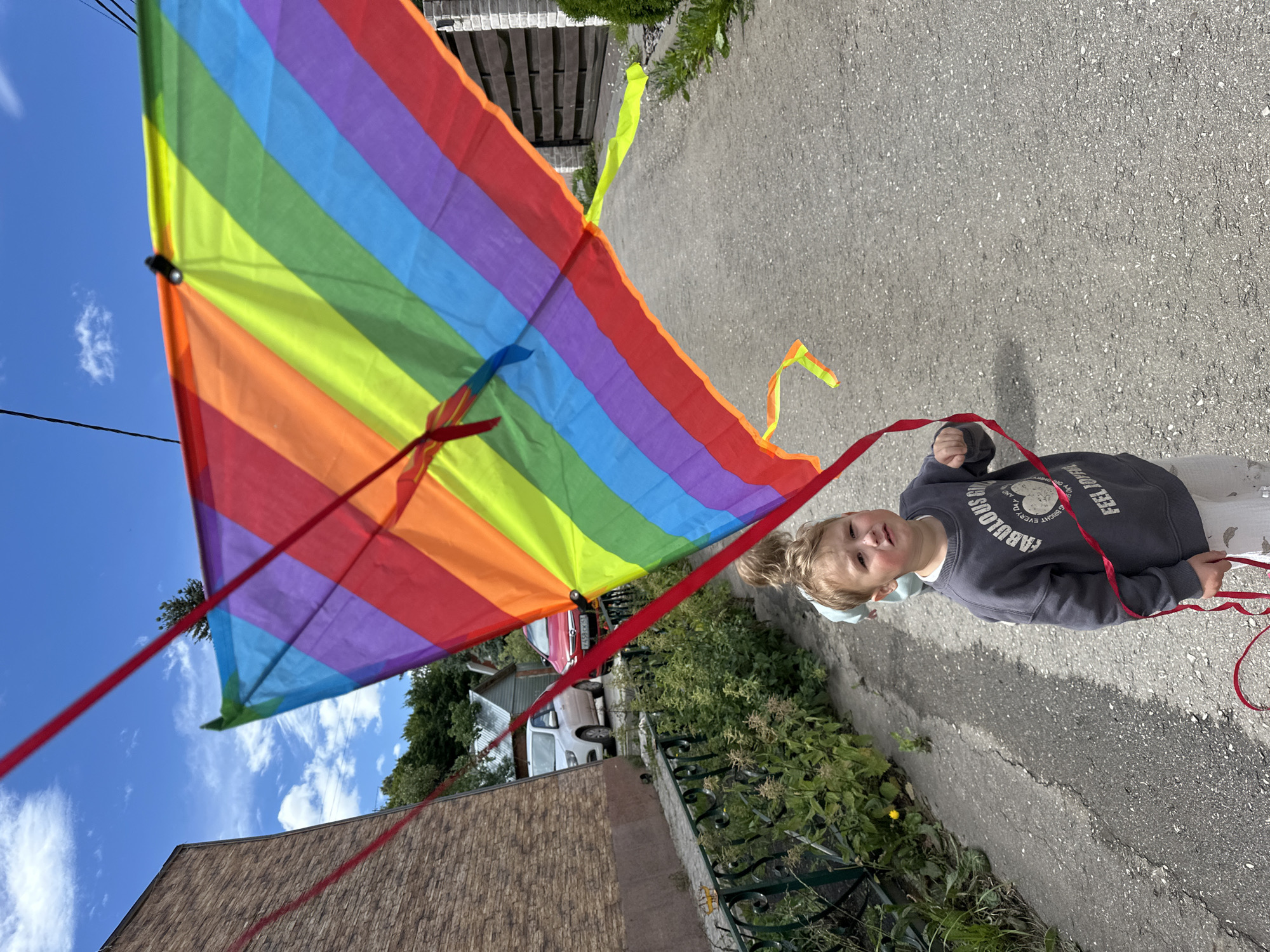 A medium shot captures a young boy, appearing to be Caucasian and under ten years old, standing on an asphalt pathway in an outdoor urban setting. He is wearing a gray crewneck sweatshirt with a heart graphic and text reading "Fabulous Girl Every Day" and "Feel Joyful", and light-colored pants. The boy is holding the string of a large, rainbow-colored kite, which dominates the upper half of the frame and extends upwards and to the right. His expression is one of mild distress or concern. Behind him, another child, partially obscured, is visible. The environment includes a brick building to the left, a fence with green foliage along the pathway, and two cars parked in the background. The sky is blue with scattered white clouds. A glimpse of a wooden fence is visible on the right side of the image. The overall scene suggests a sunny day suitable for outdoor activities. The presence of Russian license plates on the vehicles indicates the location is likely within Russia, potentially Nizhny Novgorod.
