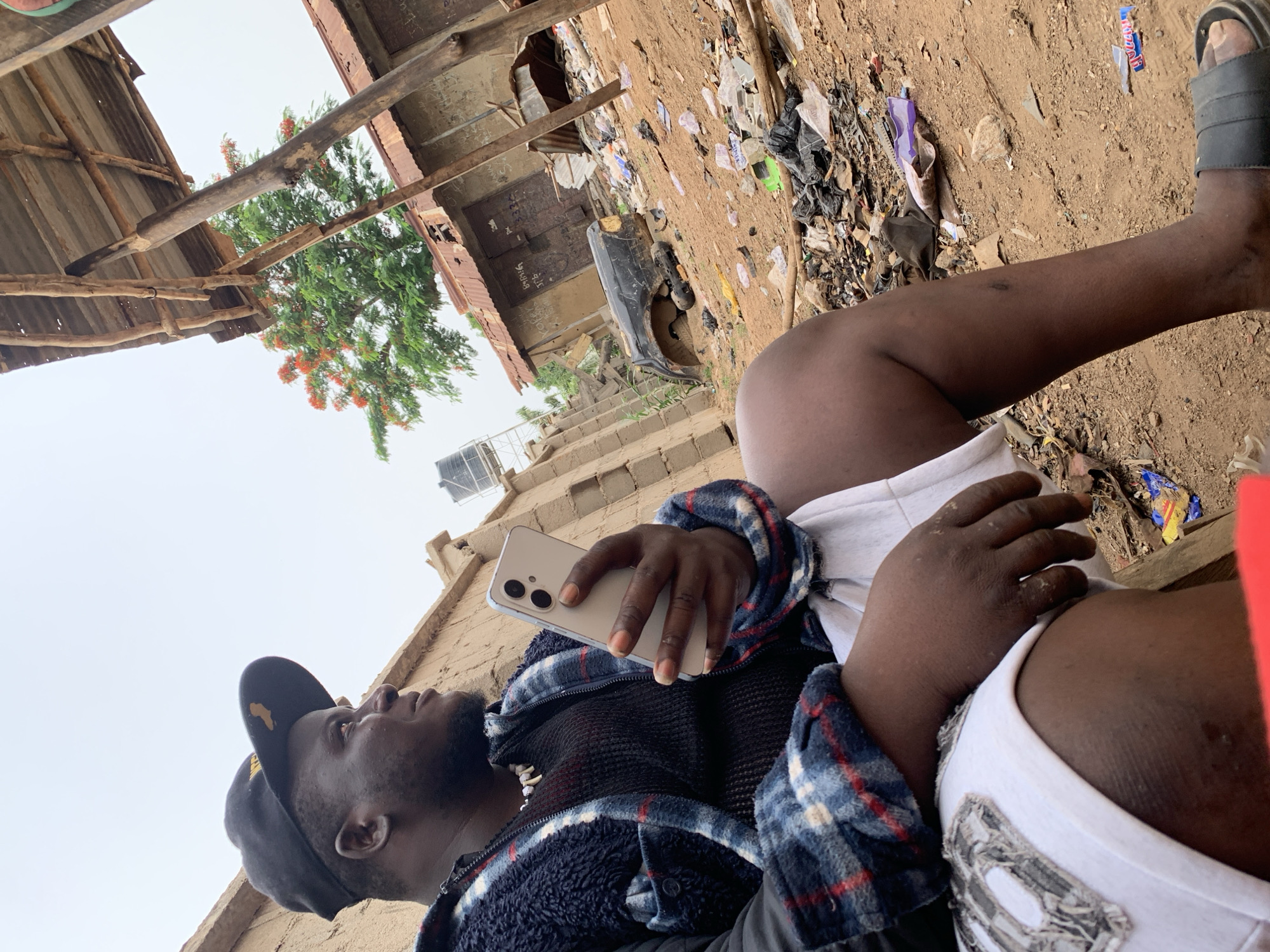 A man wearing a black baseball cap with an Africa logo, a black and blue plaid jacket, and white shorts is lying down, holding a light-colored smartphone. He is resting on his side, looking upwards, with one leg extended and a sandal on his foot. The setting appears to be a somewhat impoverished area. Debris and trash are scattered on the ground. A dilapidated wooden structure is visible to the left, and a weathered building with corrugated metal roofing is in the background. The image, if indeed located in Akwanga, Nigeria, shows possible signs of socio-economic challenges such as inadequate waste management.