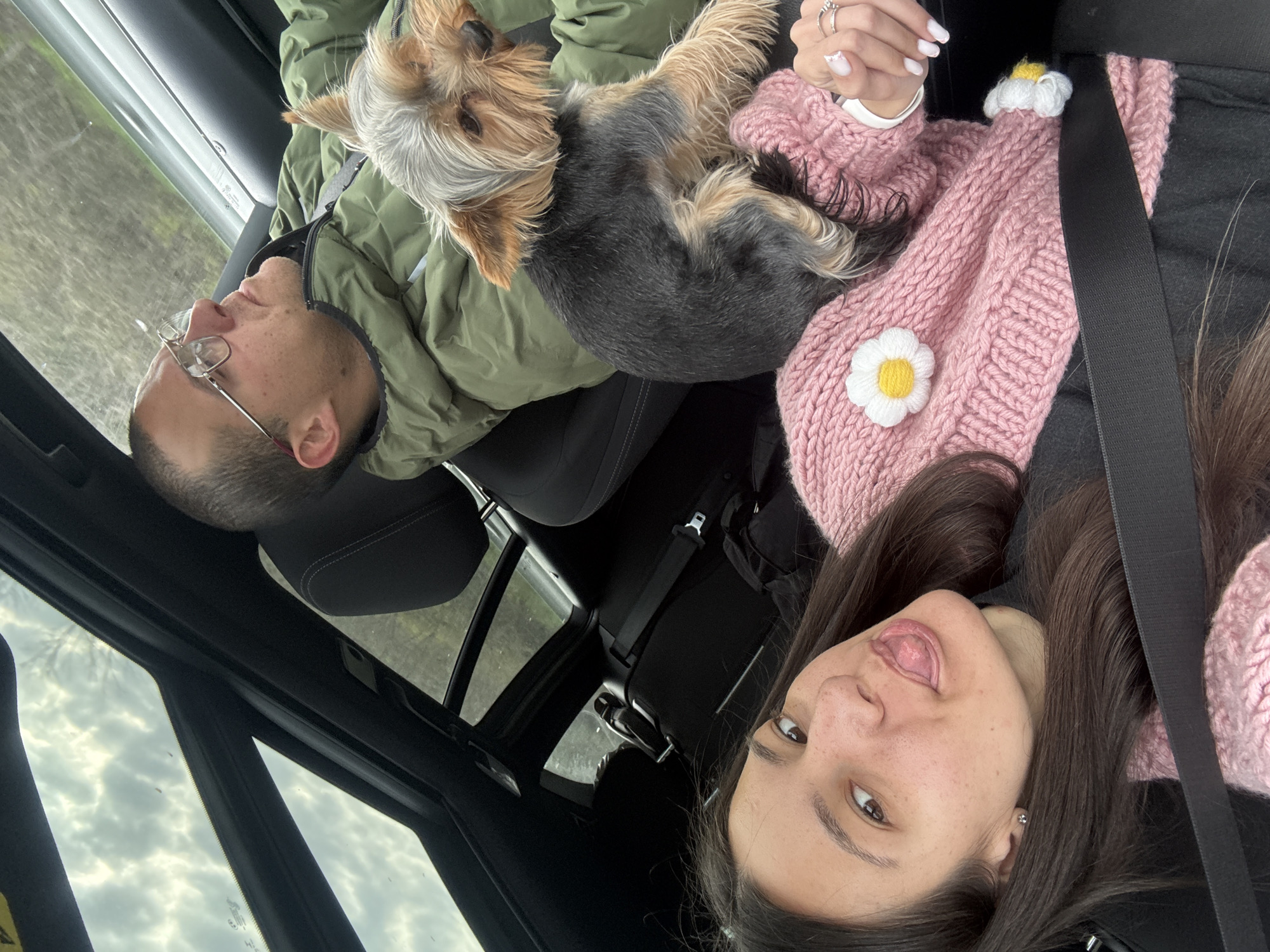 A selfie was taken inside a car showing two people and a dog. The man is on the left wearing glasses and a green jacket. A Yorkshire Terrier dog is on top of him. The woman is on the right, wearing a pink sweater with flower appliques and a seatbelt. The car has a sunroof with a view of cloudy sky. There is no evidence to suggest the location is in Kipra, Bulgaria.