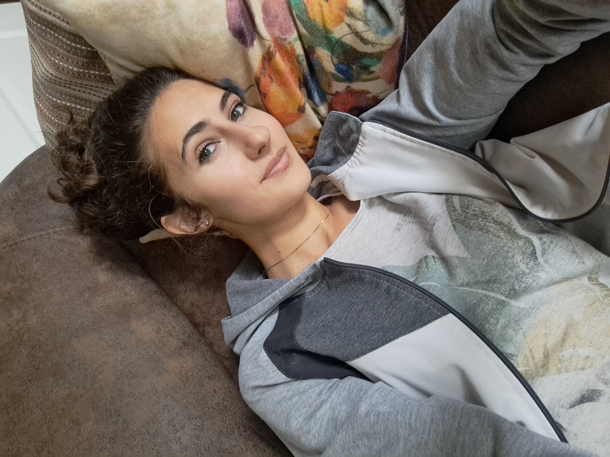 A woman is reclining on a brown upholstered sofa or couch. She is lying on her back with her head resting on a pillow with a floral design. She has curly brown hair pulled back, fair skin, and green eyes. She is wearing a light grey hooded sweatshirt with a white panel on the right side. Underneath the sweatshirt, she appears to be wearing a light grey t-shirt. A delicate gold necklace is visible around her neck. The location appears to be an indoor setting, likely a home, in an unknown city.