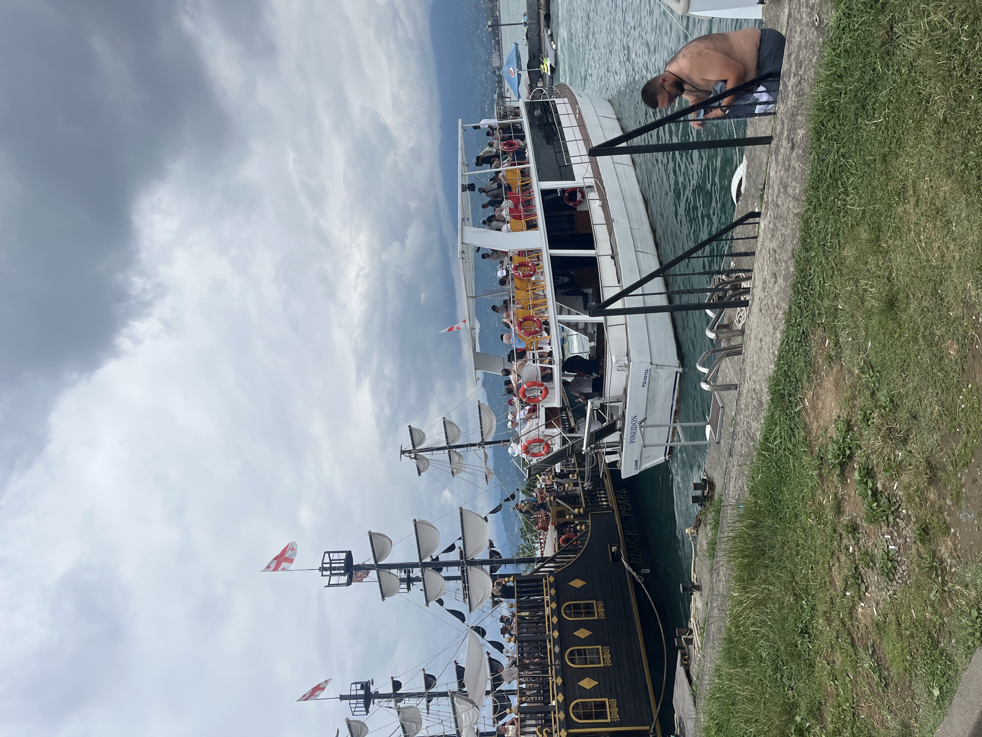 The image depicts a waterfront scene in Batumi, Georgia, featuring two boats docked side-by-side. The boat on the left is a black, pirate-themed vessel named "BLACK PEARL," adorned with white sails and flags. It appears to be carrying passengers on its decks. To its right is a white passenger boat named "POSEIDON," also with people on board. The boats are situated in a harbor with calm, turquoise water. In the foreground, a man is seated on a concrete step, appearing to be changing clothes. Behind the boats, the city of Batumi is visible with buildings and hills in the background under a cloudy sky. The overall atmosphere suggests a tourist-oriented location with recreational boat activities.