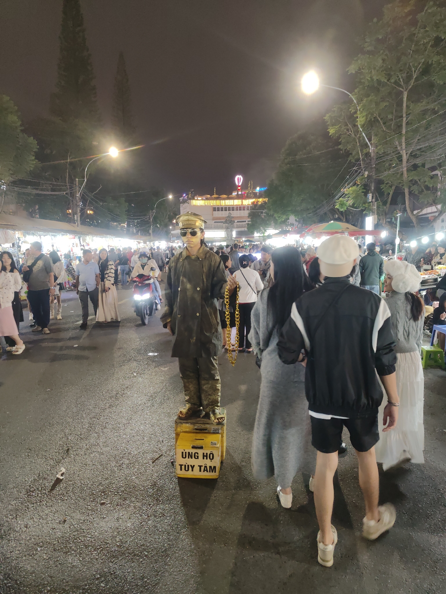 The image depicts a nighttime scene in what appears to be a busy street market or promenade in Da Lat, Vietnam. The primary subject is a person dressed as a statue, likely a street performer, painted to resemble gold and wearing a military-style uniform and sunglasses. They are standing rigidly on a yellow donation box with Vietnamese text that reads "ỦNG HỘ TÙY TÂM" (Support As You Wish).
The setting is an outdoor urban environment at night, illuminated by streetlights and the lights from numerous market stalls and buildings in the background. People are seen walking through the street, browsing stalls, and interacting with each other. Some individuals are wearing casual clothing suitable for a warm evening. A moped is visible in the mid-ground, suggesting vehicle traffic is present. Trees line the street, and in the far background, a building with bright lights, possibly a hotel or entertainment venue, can be seen.
The overall atmosphere appears lively and commercial, characteristic of a night market. The performer's pose and attire suggest an act intended to attract attention and donations. The time of day is clearly night due to the artificial lighting and dark sky. There are no overt cues about the weather, but the open-air setting suggests it is likely clear or mild. The presence of diverse individuals, market stalls, and the performer indicates a public gathering or event.