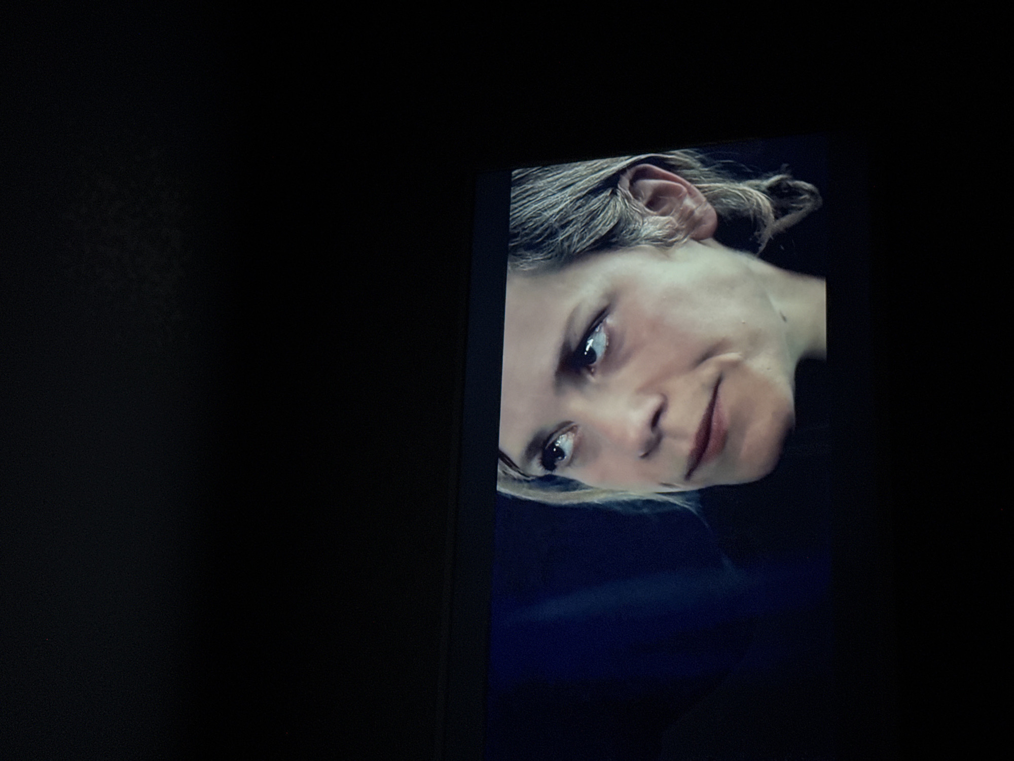 A photograph shows a headshot of a person with fair skin and short, light-colored hair, displayed on a screen within a dark environment. The person is slightly rotated, facing the viewer with a subtle, almost neutral expression. Their gaze is directed toward the left of the frame. Details such as the ear and hair texture are visible. The surrounding context suggests the image is on a television or monitor, set against a black, presumably indoor background. There is no information in the image to indicate a specific location in Mytishchi, Russia.