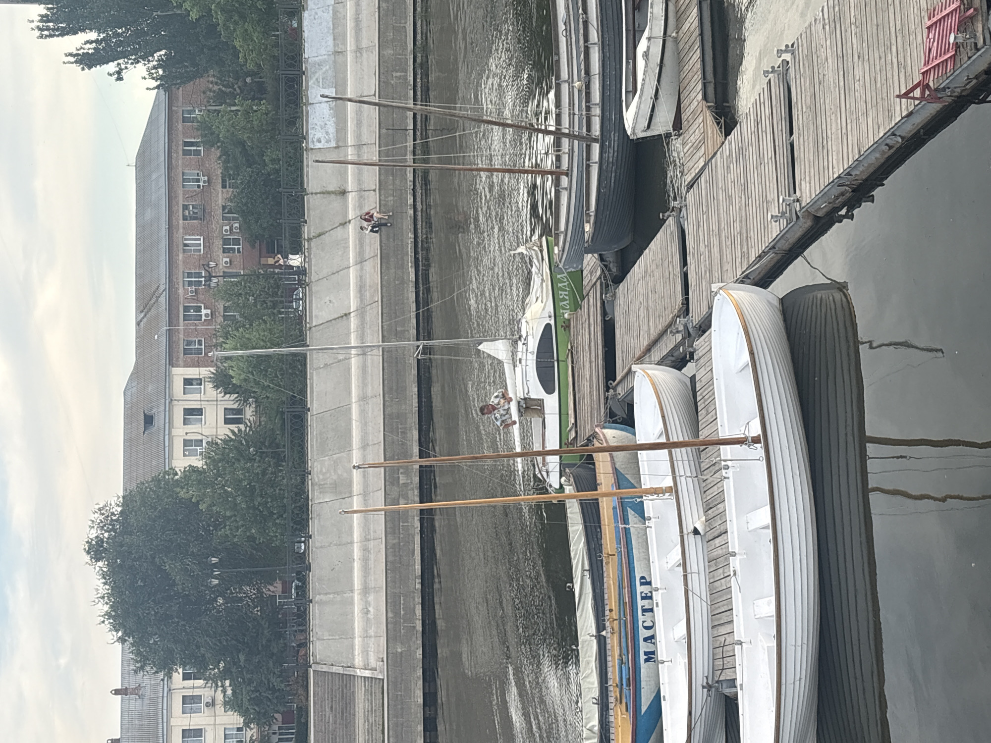 The image depicts a waterfront scene in Astrakhan, Russia, likely along the Volga River or one of its canals. In the foreground, several white rowboats are docked at a wooden pier. One of these boats has "МАСТЕР" written in blue letters on its side. To the right, a darker-colored boat is also moored. In the middle ground, a man in a patterned shirt stands on the deck of a green and white sailboat. Further back, a wider concrete embankment lines the water, with two people seated on its edge. Behind the embankment, buildings with multiple windows and tiled roofs are visible, suggesting an urban setting. Trees and vegetation are interspersed between the buildings and along the embankment. The sky is overcast with diffused light.