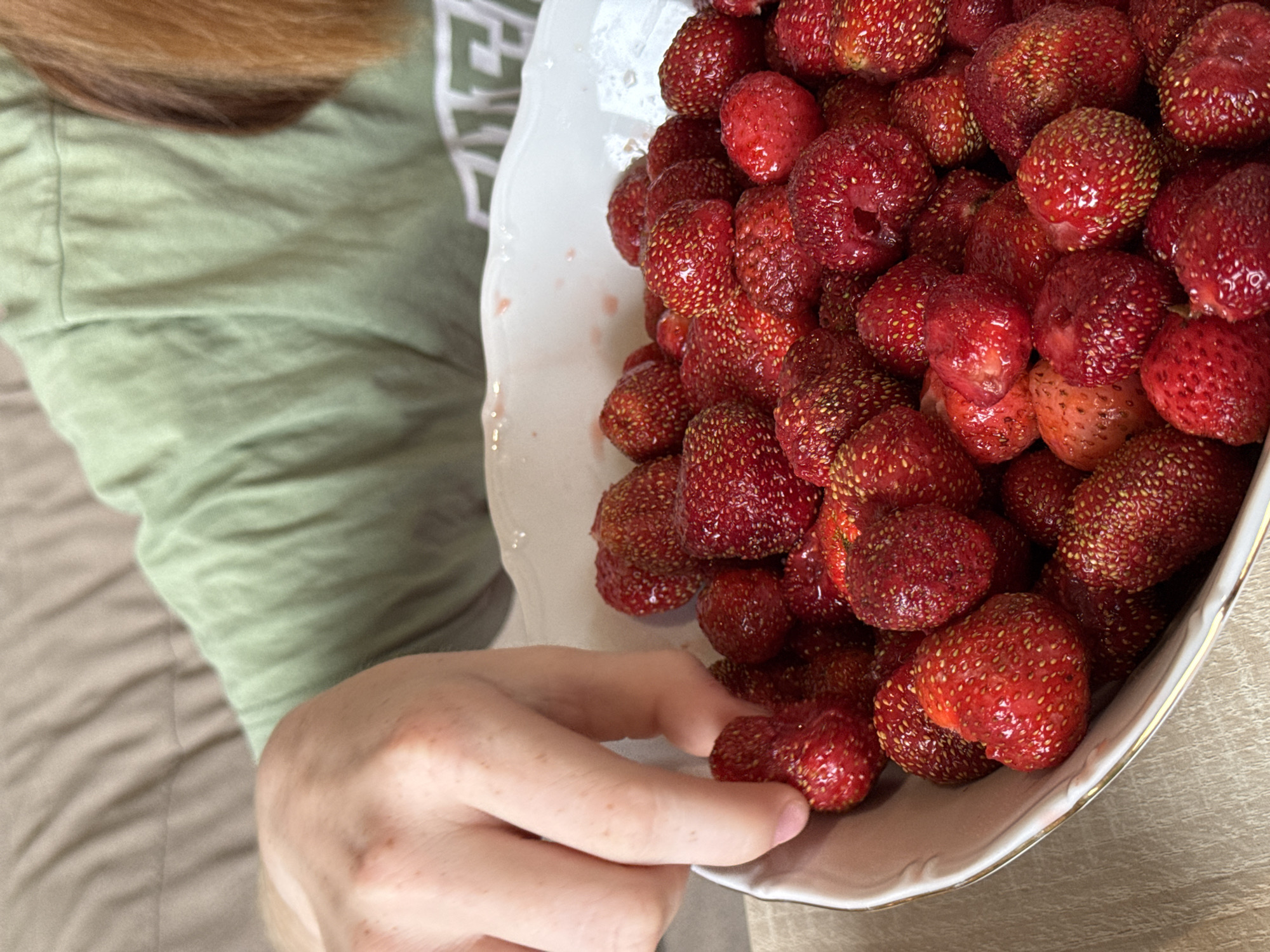 A person's hand is reaching into a white bowl filled with fresh strawberries. The bowl has a gold trim. The person is wearing a light green t-shirt. The background shows a couch with a textured fabric. The scene appears to be indoors. There is no indication of the city of Chaikovsky, Russia.