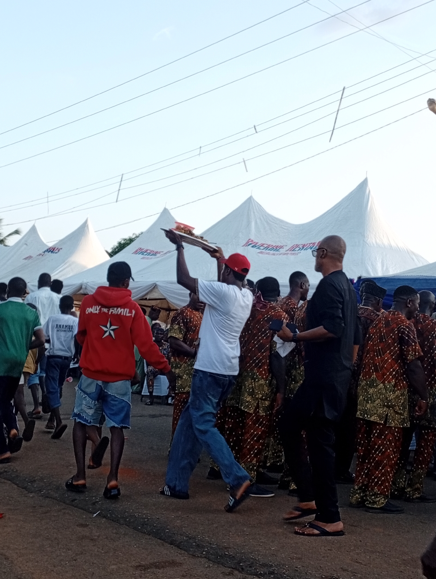 A medium-wide shot captures an outdoor gathering in Nsugbe, Nigeria, with several white tents forming a backdrop. In the foreground, a man in a red hooded sweatshirt with the text "ONLY THE FAMILY" and a white star emblem is captured from behind as he walks away from the camera. He is wearing distressed denim shorts and dark sandals. To his left, a man in a white t-shirt and red cap is lifting a tray with food items. To the right of the man in the red hoodie, a bald man with glasses in a black shirt and black pants is looking to his left. A crowd of people dressed in matching brown and green patterned clothing are visible, some interacting with each other and others walking. The ground is paved with asphalt. The sky is light blue with faint clouds.