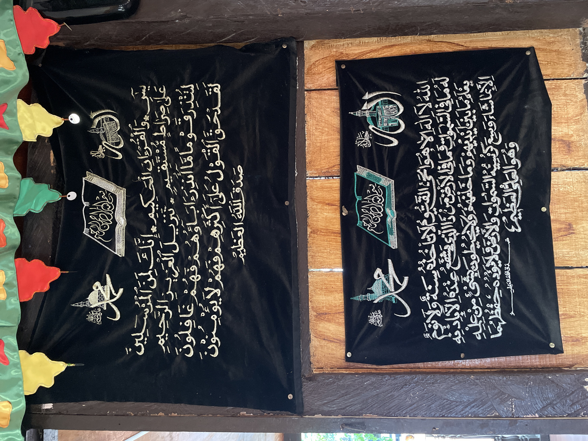 The image displays two black banners with Arabic calligraphy and Islamic imagery, hung on wooden walls. Both banners appear to be religious in nature, likely religious verses or declarations. The upper banner features an image of an open book and a mosque or religious structure. The lower banner also contains calligraphy, and features an image of a mosque and a depiction of Allah in Arabic script. The banners are adorned with colorful fabric streamers at the top. The scene is indoors, with natural lighting suggesting it might be during the day. The wooden elements of the walls suggest a rustic or traditional setting. Based on available information, this scene is located at the Grand Mosque of Buhangin in Davao City, Philippines.