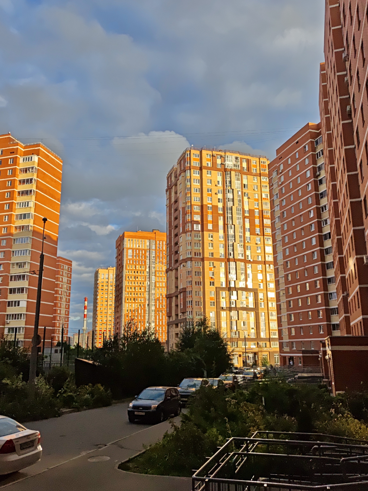 In Shcherbinka, Russia, the sun casts a warm glow on a row of towering, modern apartment buildings. The buildings, predominantly orange and red brick with white trim, stand tall against a dramatic sky filled with a mix of blue and dark, moody clouds. The architecture is characterized by repetitive window patterns and balconies, common in urban residential complexes.
In the foreground, a paved road curves gently, bordered by lush green trees and bushes that add a touch of nature to the urban landscape. A few cars are parked along the roadside, including a dark-colored sedan and a lighter-colored vehicle further down. One car, a black sedan, is clearly visible on the right side of the road. The scene suggests a late afternoon or early evening, with the low angle of the sun creating strong highlights and shadows on the buildings. In the distance, a tall, striped industrial chimney punctuates the skyline, hinting at the industrial or utility infrastructure surrounding the residential area. The overall impression is one of a contemporary urban environment, with a blend of new construction and managed greenery.
