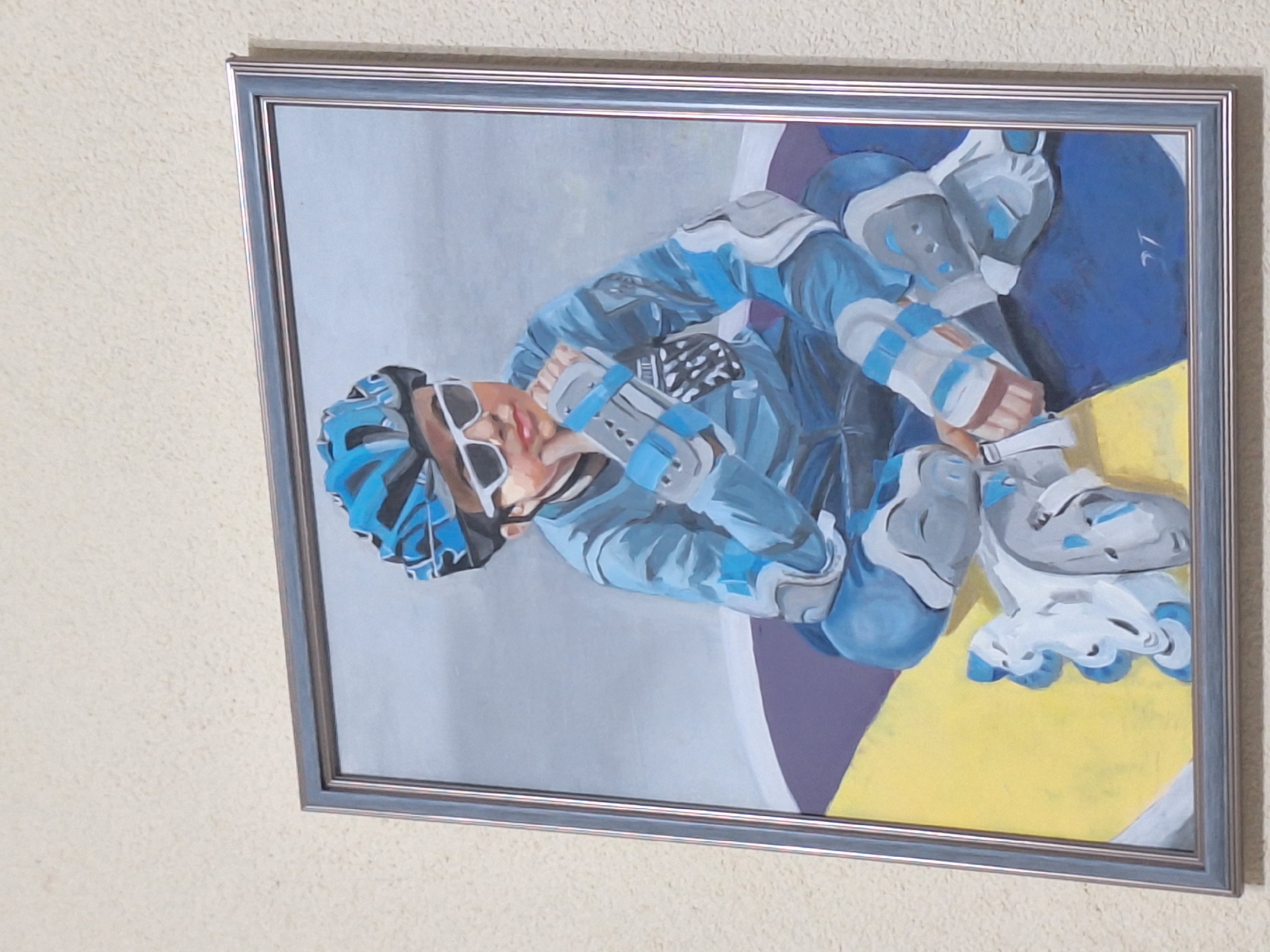 The image shows a framed painting of a person in roller skating gear hanging on a light-colored wall. The person is wearing a blue helmet, sunglasses, and protective pads. They are seated, adjusting their roller skates. The background of the painting consists of gray, purple, and yellow sections. The frame is silver. There is no indication of the painting's location being in Varna, Bulgaria.