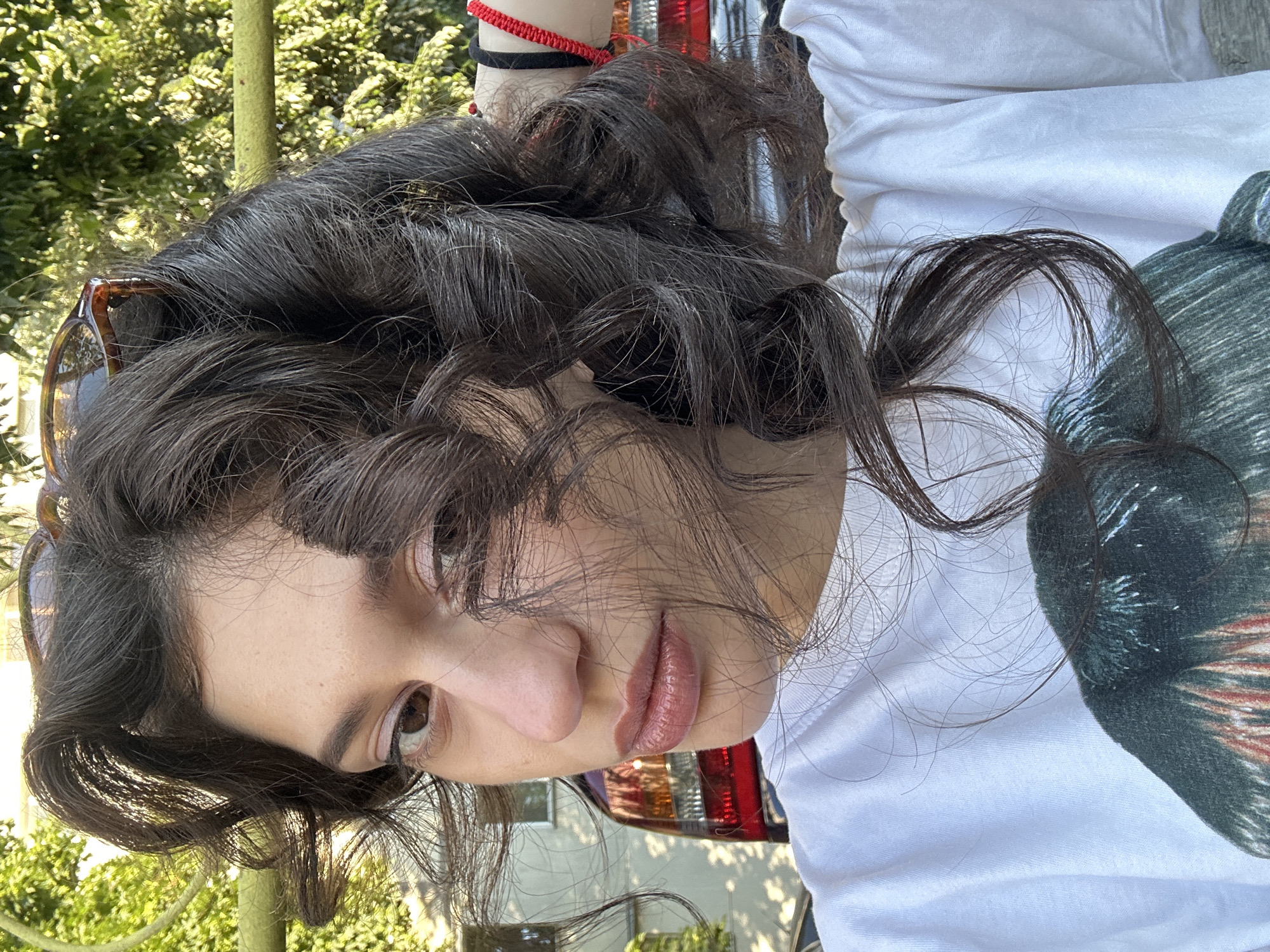 A young woman with dark, curly hair is pictured outdoors, with a blurred background suggesting a residential area or park in Sofia, Bulgaria. She is wearing a white t-shirt with a graphic print. Her hair is partially obscuring her face, and she is looking slightly away from the camera. In the background, a portion of a car's taillight is visible, along with greenery and what appears to be a building. The lighting suggests it is daytime.