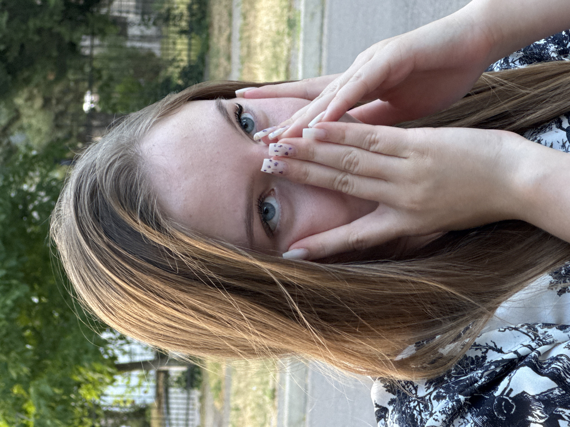 A young Caucasian female with long, light brown hair and blue eyes is captured in a close-up shot, her face partially obscured by her hands. She is wearing a black and white patterned top. Her fingernails are long and white, with some featuring a delicate purple floral design. The background suggests an outdoor urban or suburban setting, with trees and a fence visible, consistent with locations in Tiraspol, Moldova, such as the area near the Dniester River or residential streets with greenery. The lighting indicates it is daytime, likely with natural sunlight.