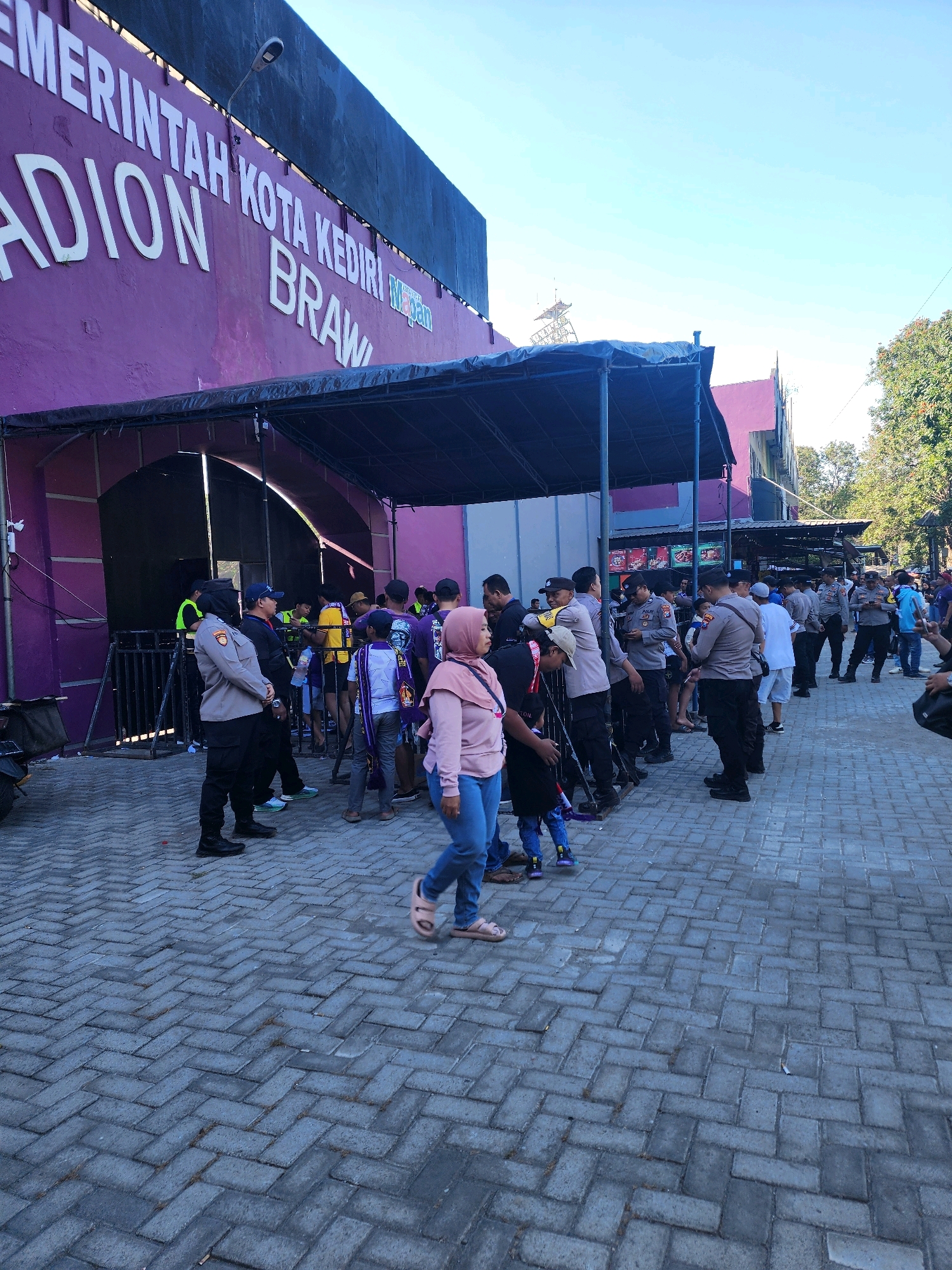 The image shows the exterior of a stadium in the city of Kediri, Indonesia. The stadium has a large purple facade with white lettering that reads "PEMERINTAH KOTA KEDIRI STADION BRAWIJAYA". A crowd of people, including security personnel in uniform, are gathered outside the stadium entrance. The ground is paved with grey interlocking bricks. Some individuals appear to be queuing to enter the stadium, while others are standing around. A black motorcycle is partially visible on the left side of the frame. The sky is clear and blue.