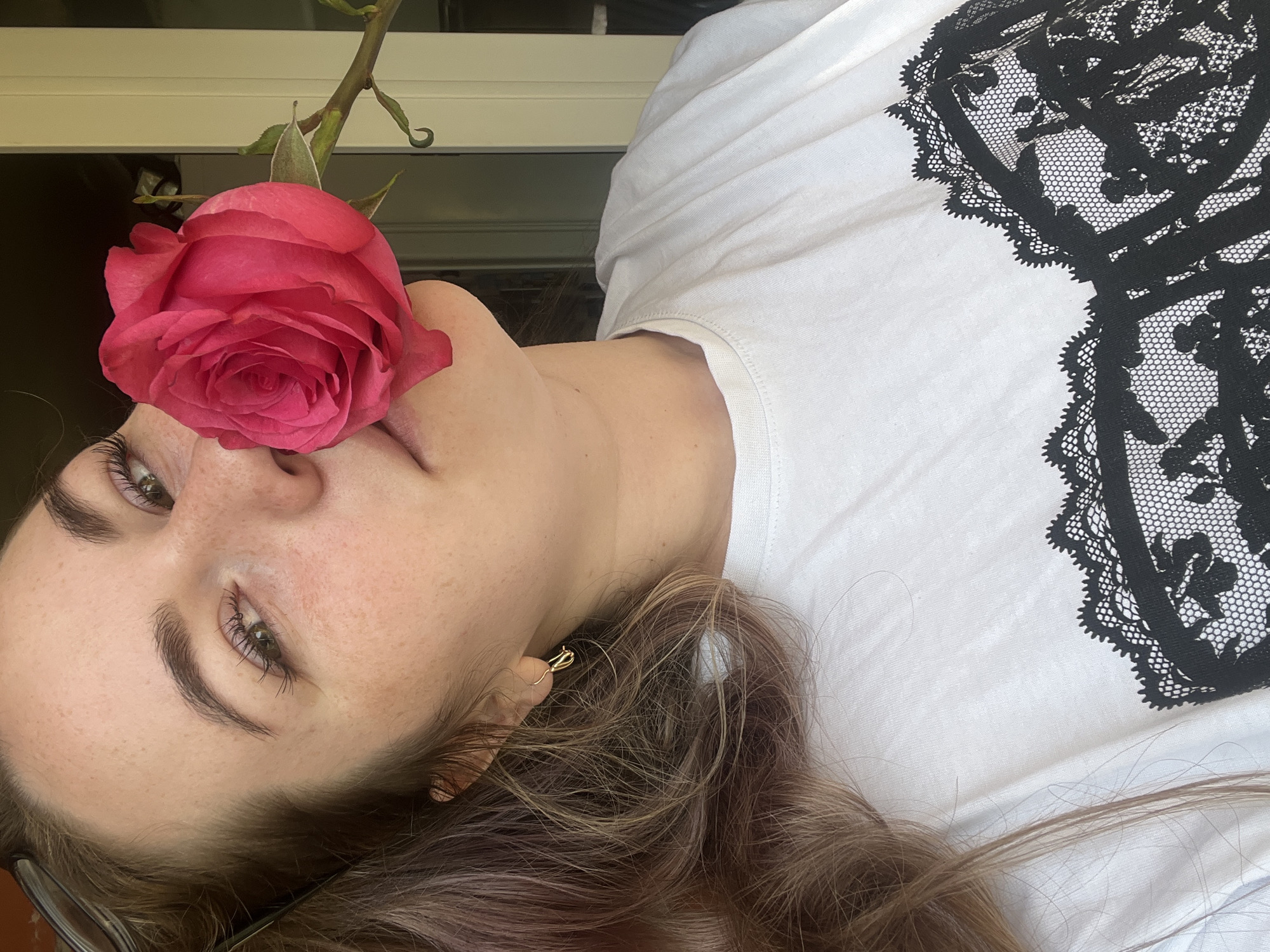 A woman with fair skin and brown hair poses with a pink rose held against her nose. She is wearing a white t-shirt with a black lace design on the upper right side. She has light green eyes and freckles. Her head is tilted to the side. A gold earring is visible in her left ear. A window is partially visible in the upper left corner. The location is unknown and is not necessarily in the city of Chaikovsky, Russia.