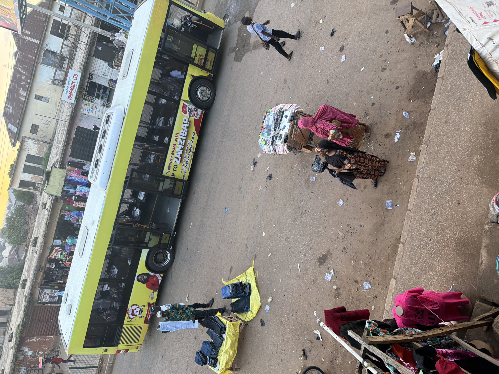 The image captures a street scene in Bukuru, Nigeria. A bright yellow bus with advertising is parked on the right side of the road. In the foreground, a vendor is displaying folded jeans on a table covered with a yellow tarp. Several people are walking or standing in the street, some carrying items. The street is paved with asphalt and appears to be a busy marketplace, with shops and buildings lining the background. Debris is scattered on the ground, indicating an active urban environment.