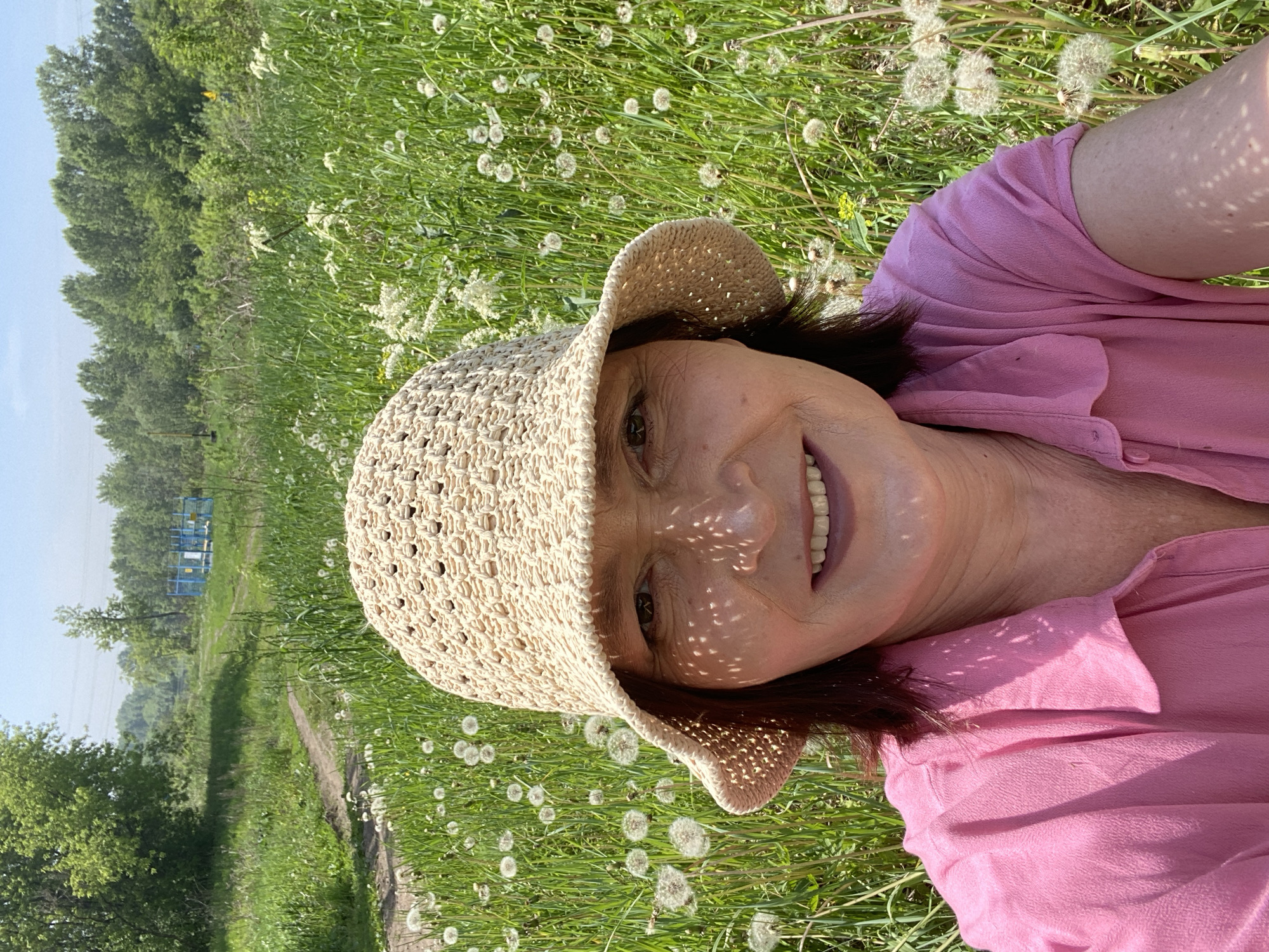 An outdoor, eye-level selfie-style photograph depicts a smiling, middle-aged woman positioned in a lush green field under bright daylight in Novokuznetsk, Russia. The woman, with dark brown hair, wears a light-colored, loosely woven or crocheted straw bucket hat that casts distinct dappled shadows across her forehead and cheeks. She is dressed in a collared, short-sleeved pink shirt. Her expression is a broad smile, revealing upper and lower teeth, and her eyes are directed towards the camera.
The background consists of an expansive field of vibrant green grass, heavily interspersed with numerous white dandelion puffballs. A dirt path is visible curving through the grass in the mid-ground on the left side of the frame. Beyond the immediate field, a dense line of green deciduous trees forms the horizon, with a partial view of what appears to be a body of water or a distant cleared area in the far background. A blue, possibly metallic, structure with multiple uprights is situated in the mid-ground to the right of the path, partially obscured by foliage. The sky above is clear and light blue. Sunlight illuminates the scene from an elevated position, creating prominent shadows and highlighting textures.