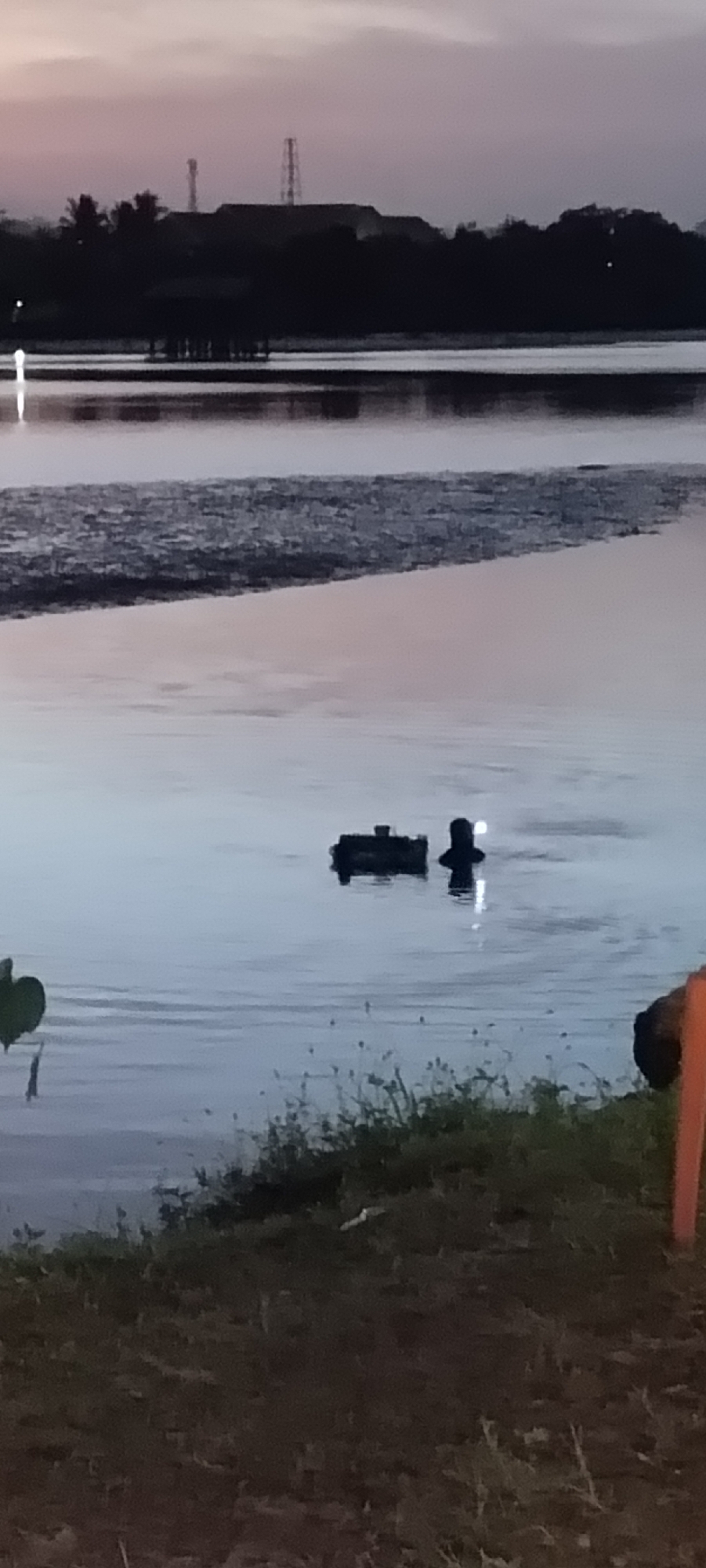 The image shows a dimly lit scene at dusk, likely near a body of water. In the foreground, there is a grassy area with some vegetation. The middle ground features a large body of water, possibly a lake or ocean, with a hazy line of trees and structures in the background.
In the water, there are two distinct objects. One is a dark, rectangular object, possibly a buoy or a piece of debris. Near it, a person is visible, with their head and shoulders above the water, appearing to be swimming or wading. The person is silhouetted against the water's surface.
The sky above is a pale purple and gray, indicating the transition from day to night. The overall atmosphere is calm and quiet. Given the location mentioned, this scene could be a waterfront area in Lhokseumawe, Indonesia, perhaps a reservoir or coastal inlet, captured during twilight.
