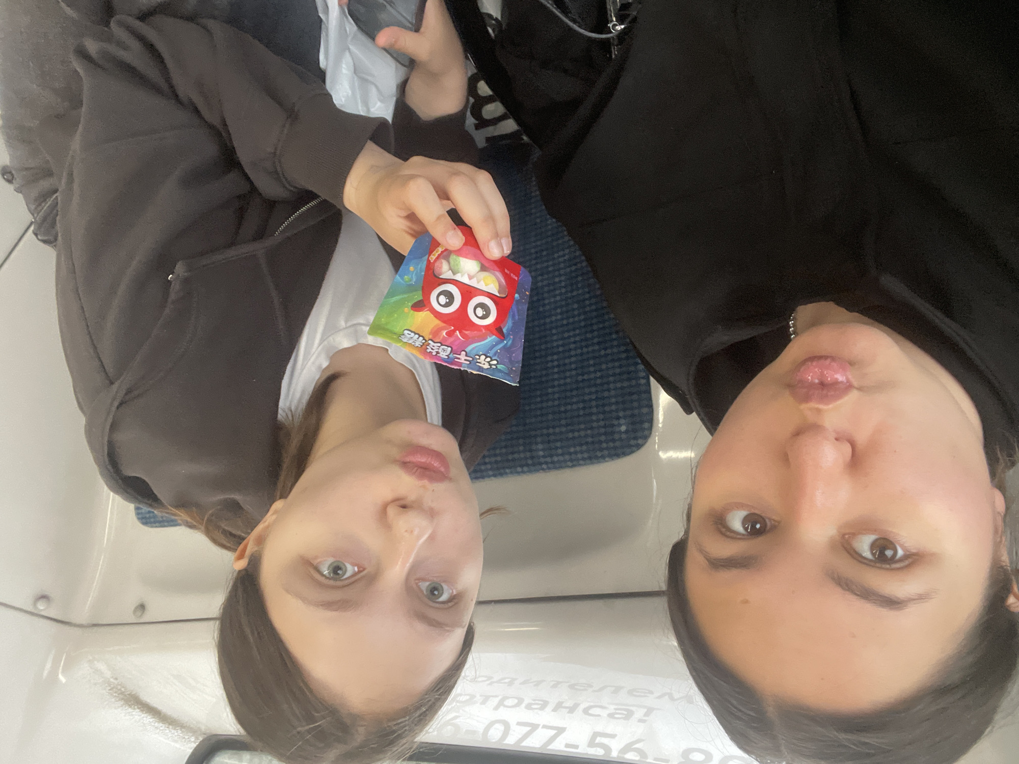 Two young women are seated on a train. The woman on the left is facing forward, looking at the camera with her lips pursed. The woman on the right is turned towards the left, holding a colorful candy packet with a cartoon monster on it. Both women are wearing casual clothing, and appear to be in a train car. The background shows seats and the interior of the train car. There is text visible on the wall behind them, which appears to be a train schedule or advertisement, possibly indicating they are in or near Рассудово, Russia.