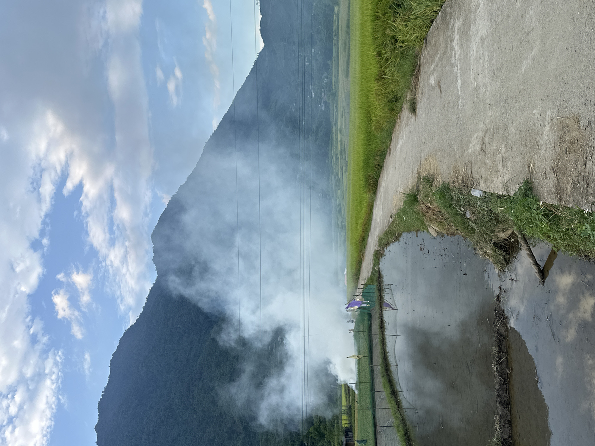 This image depicts a rural mountainous landscape in Gia Hội, Vietnam, characterized by agricultural activity. In the background, large, dark green, forested mountains rise steeply under a partly cloudy blue sky. Scattered white and grey clouds are visible across the sky.
A dense, white-grey smoke plume billows from the mid-ground on the left, extending upwards and to the right, partially obscuring the lower slopes of the mountains and several horizontal utility lines running across the scene. Near the origin of the smoke, two individuals are faintly visible, one wearing a conical hat, suggesting human activity related to the burning. Adjacent to the smoke, in a cultivated field, two figures resembling scarecrows, one with yellow fabric and another with purple, are positioned.
The mid-ground features vibrant green agricultural fields, likely rice paddies, extending towards the base of the mountains on the right. In the foreground, a narrow, unpaved concrete path curves slightly from the lower center towards the mid-ground, separating the green fields on the right from water-filled cultivated sections on the left. These water-filled areas reflect the sky and clouds, with a visible earthen divider separating two sections. Green netting or fencing encloses parts of these water-filled fields. A white plastic bottle lies on the edge of the path in the foreground, and a wooden branch is partially submerged in the water on the lower right. The scene is illuminated by daylight.