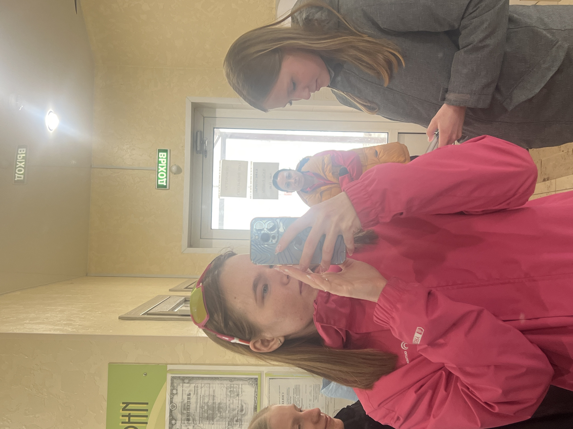 The image shows two young women indoors in Gory, Russia. One woman is holding a smartphone and taking a picture of the other woman. The first woman is wearing a pink jacket and has sunglasses on her head. The second woman is wearing a gray jacket. The background includes a hallway with signs in Russian, a door, and other people in the distance. The sign on the left reads "ВЫХОД," meaning "EXIT," and the sign on the right reads "ВХОД," meaning "ENTRANCE."