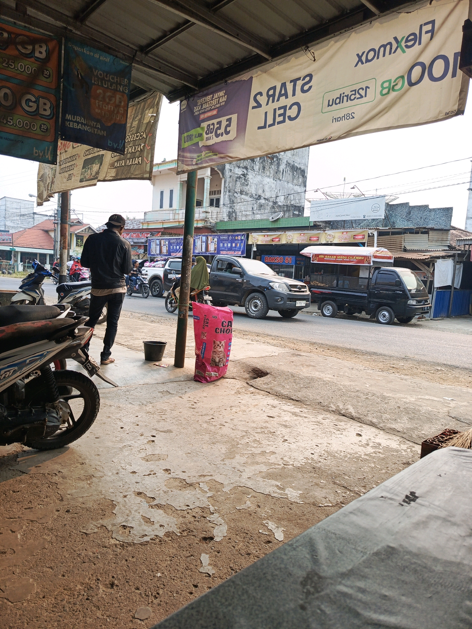 The image depicts a street scene in front of a small shop or roadside stall in Megang Sakti II, Indonesia. In the foreground, a person wearing a black jacket and dark jeans stands facing away from the camera, looking towards the street. A black bucket and a large pink bag with cat images and text are on the sidewalk to the right of the person. Several motorcycles are parked on the left side of the image. In the background, various signs and banners hang overhead, advertising mobile data plans and other services. A black pickup truck and a white pickup truck are visible on the street, along with other vehicles and pedestrians. The environment appears to be a commercial area with buildings lining the street. The lighting suggests it is daytime, possibly late afternoon, given the long shadows.