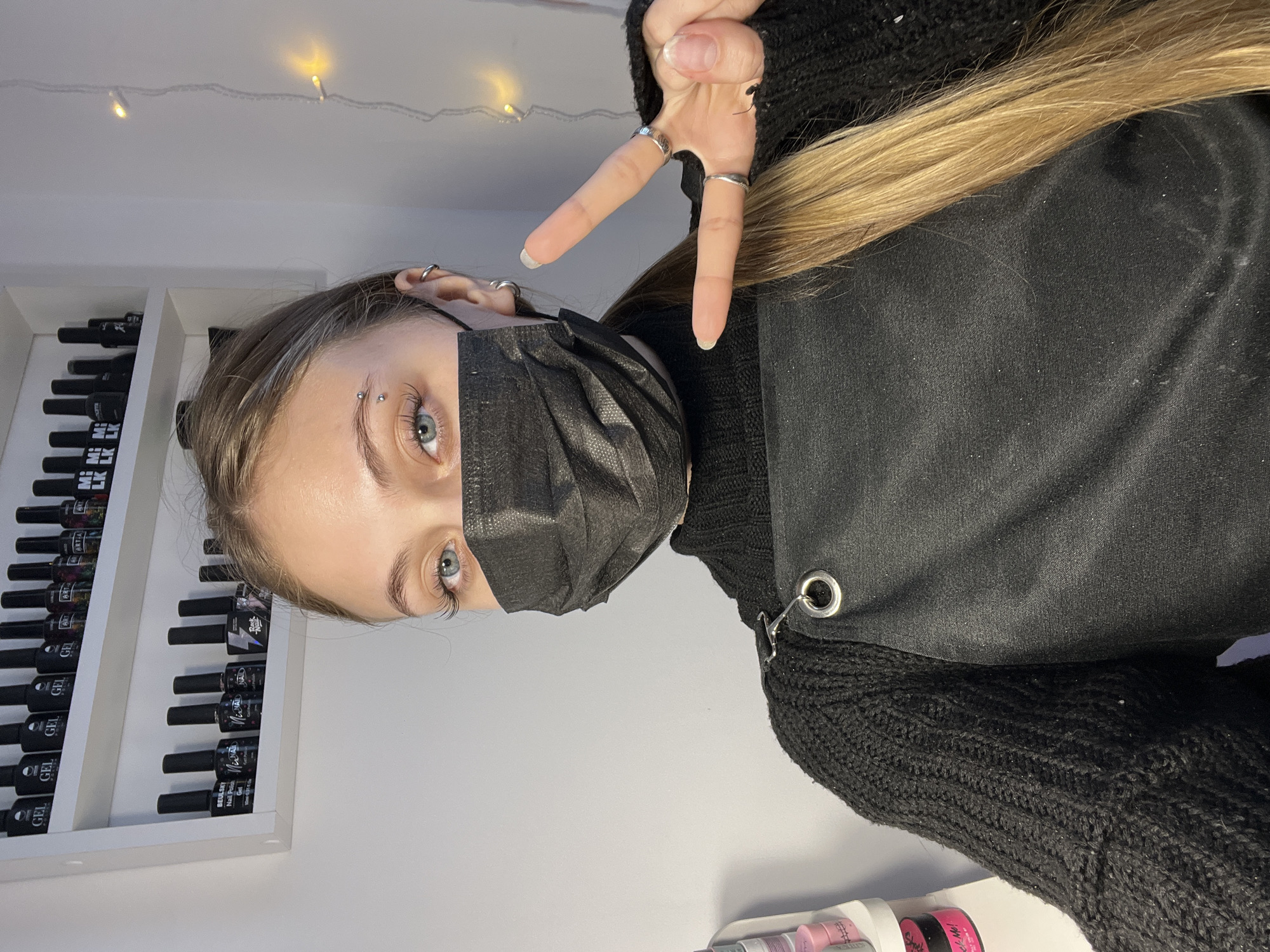 A person wearing a black face mask and making a peace sign with their fingers is the primary subject of this indoor photograph. The individual has an eyebrow piercing and their light brown hair is visible. They are wearing a black sweater and a black apron or garment with a metal ring detail.
To the left, there is a white shelf unit filled with black nail polish bottles, each with a small label. Above the shelf, a string of lights is faintly visible. A small container with pink products is partially visible in the lower right corner of the image. The wall behind the subject is a plain, light color.