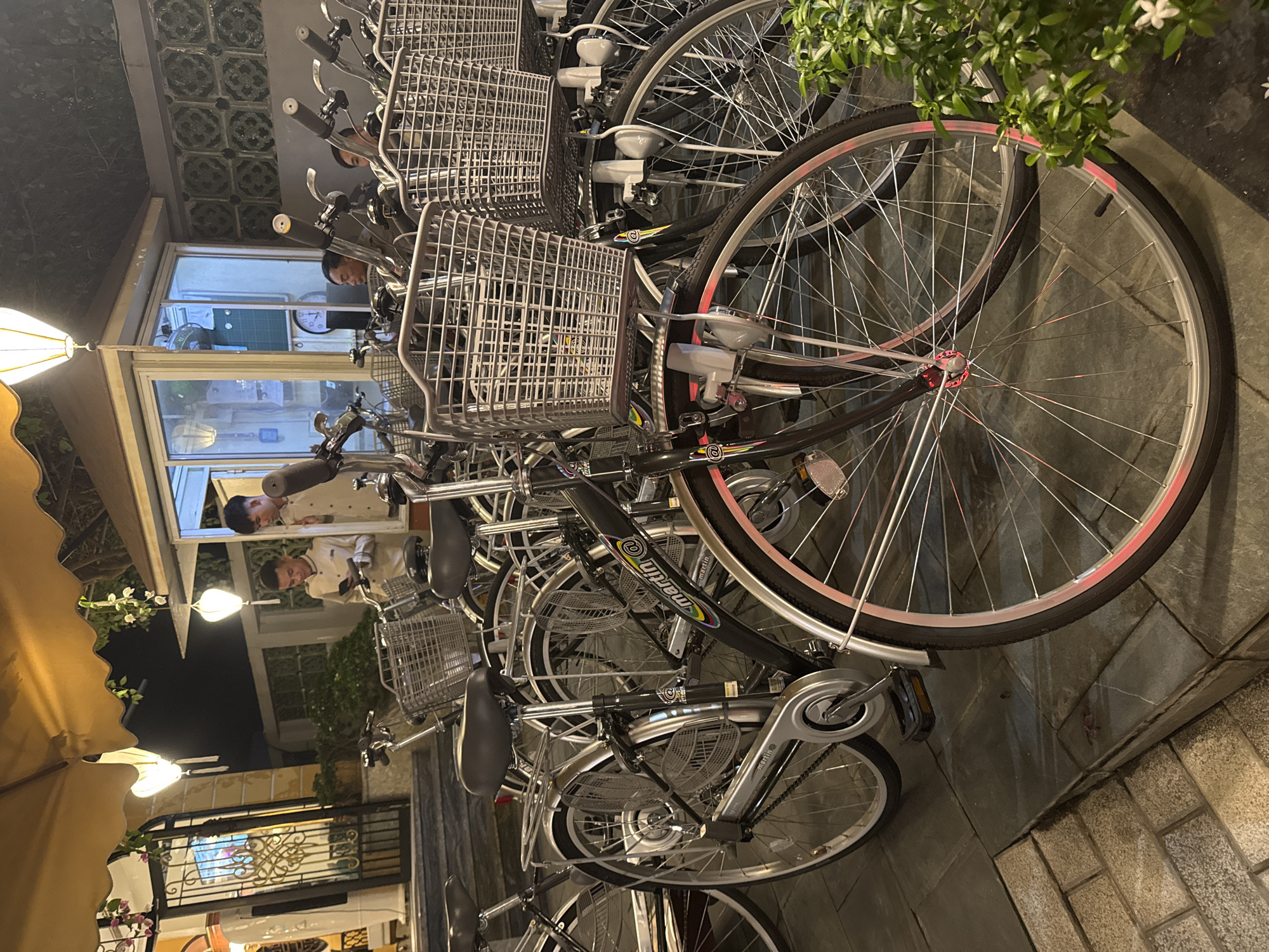 The image shows a row of parked bicycles in Hội An Ward, Vietnam. The bicycles are mostly silver and black with wire baskets on the front. Some have lights attached to the handlebars. The tires of the bicycle in the foreground has a red stripe. In the background, two people wearing white shirts are visible through a window, suggesting the setting is near a business or restaurant. A yellow awning and outdoor lighting are also visible. The bikes are parked on a tiled pavement.