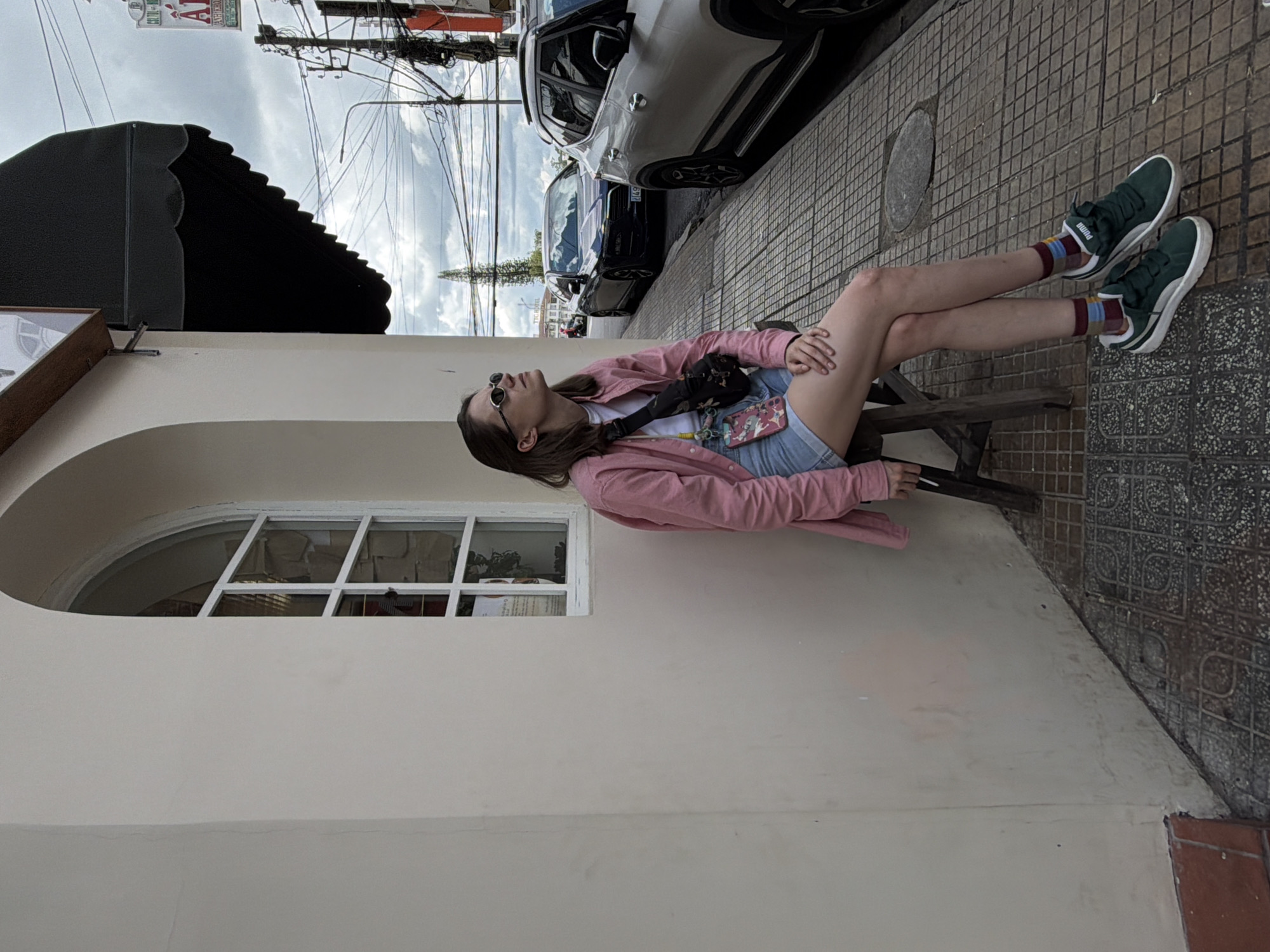 A young woman is sitting on a dark wooden stool on a sidewalk in Da Lat, Vietnam. She is wearing a pink button-down shirt, denim shorts, a white t-shirt, sunglasses, and dark green Puma sneakers with white accents and striped socks. She has a black fanny pack across her chest with a pink patterned phone case hanging from it. She is looking up and to her left, and her right hand is resting on her knee. The sidewalk is paved with a mosaic-like pattern of gray and white tiles. Behind her, there is a light-colored building with an arched window. The street is lined with cars, including a silver SUV on the right and a dark sedan further down. Electrical wires and a lamppost are visible in the background, as well as a tall, slender tree. The sky is overcast with clouds.