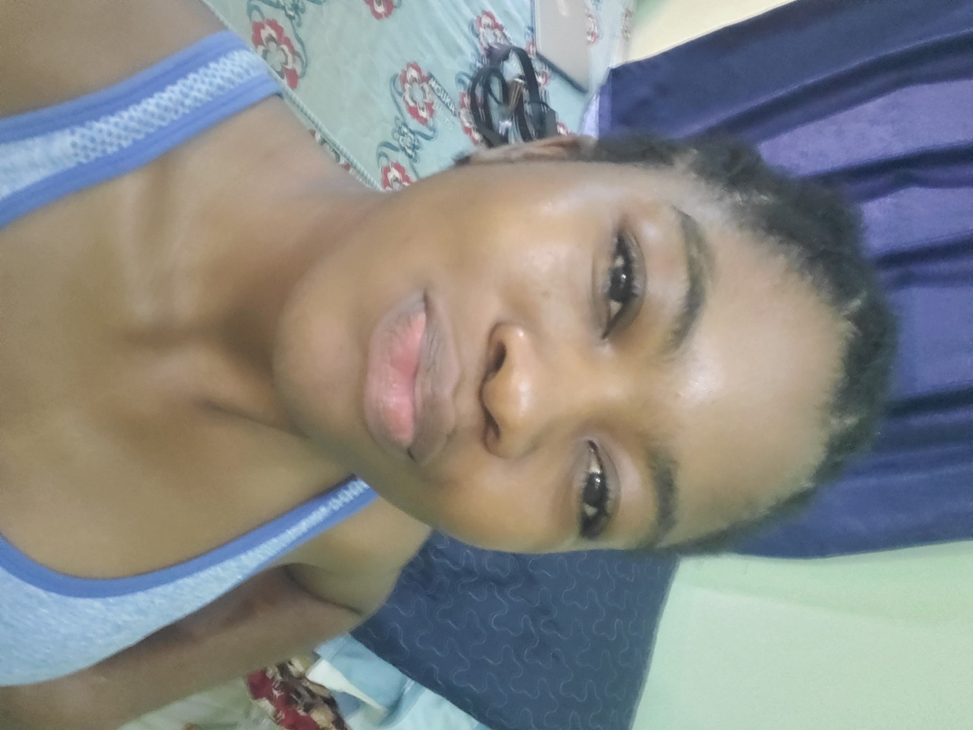 A medium-close shot depicts a woman with dark skin and short, dark hair lying on her back. She is wearing a light blue tank top. Her gaze is directed towards the camera. Behind her is a blue pillow and a bedspread with a floral pattern and a pair of glasses. The location, Barden Yana, Nigeria, can not be determined from the image.