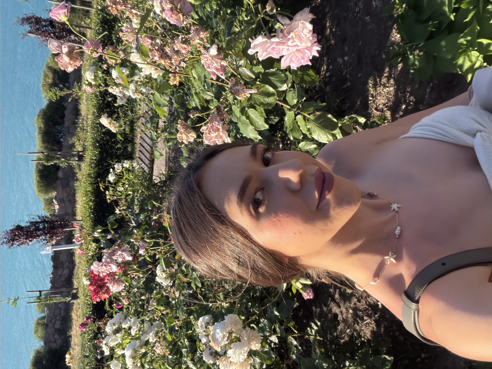 In Sozopol, Bulgaria, a young woman with brown hair is captured in a selfie. She is positioned in the foreground, with her right shoulder and face visible. She is wearing a white top and a necklace with seashell pendants. Behind her, a lush rose garden blooms with various colored roses, including white, pink, and red. The garden is bordered by a stone wall and a hedge, with the blue sea and sky visible in the background. The lighting suggests it is daytime, possibly late afternoon due to the warm sunlight illuminating the scene.