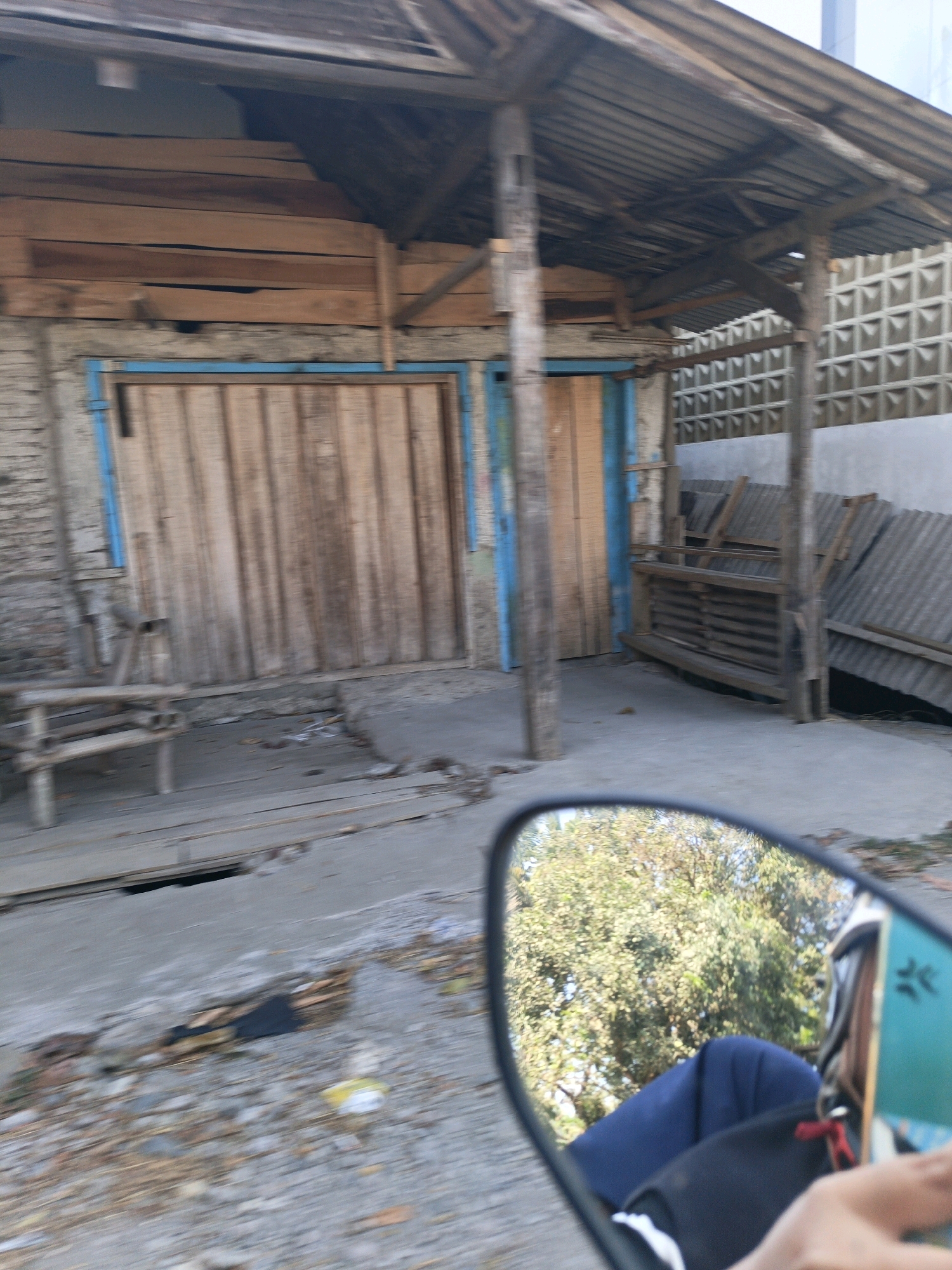 The image captures a street scene in Sidodadi, Indonesia, focusing on a weathered, open-air structure with a corrugated metal roof supported by wooden beams. The structure appears to be a shop or workshop. In the foreground, a motorcycle side mirror reflects trees and a person's arm holding a turquoise phone. The building's walls are a mix of brick and what appears to be wooden planks, with large, rustic wooden doors. A simple wooden bench is positioned to the left of the doors. To the right, stacks of corrugated metal roofing sheets lean against the wall. The ground is a mix of concrete and dirt, with scattered debris. The overall impression is one of a humble, utilitarian urban or semi-urban environment.