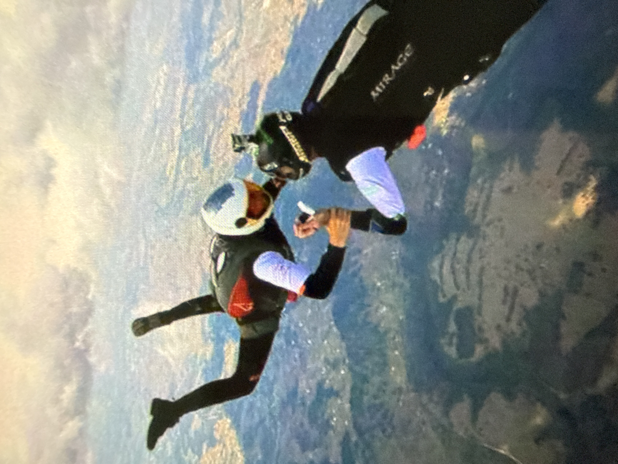 Two skydivers are in freefall. One skydiver is wearing a white, blue, and orange helmet, a black jumpsuit with red accents, and light-colored sleeves. The other skydiver is wearing a black jumpsuit with the word "MIRAGE" on it, a helmet with a camera attached, and light-colored sleeves. The background shows a landscape with patches of light and dark areas, possibly indicating terrain features or cloud cover. The location is in the city of Unknown, Unknown.