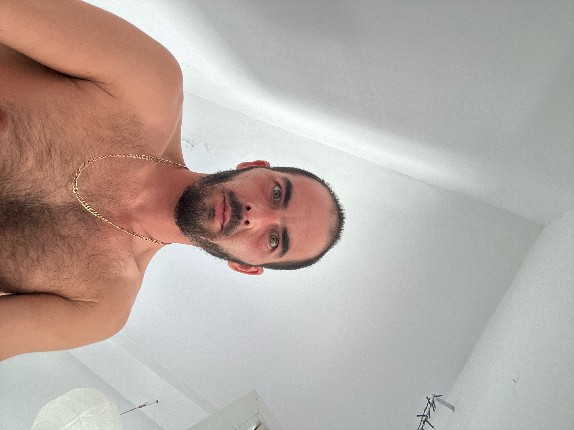 The image shows a low-angle, close-up, portrait-style shot of a man from the chest up, appearing to be looking directly at the camera. The man has short dark hair, a beard, and is wearing a gold chain. He is shirtless, revealing chest and shoulder hair. The background is a plain, white, textured ceiling, with a hint of a white paper lantern and some string lights visible in the lower portion of the frame. The lighting is bright and diffused, suggesting an indoor environment. There are no discernible elements in the image that would place the scene specifically in Budva, Montenegro, or any other city. The perspective suggests the photographer is positioned directly above the subject, looking down.